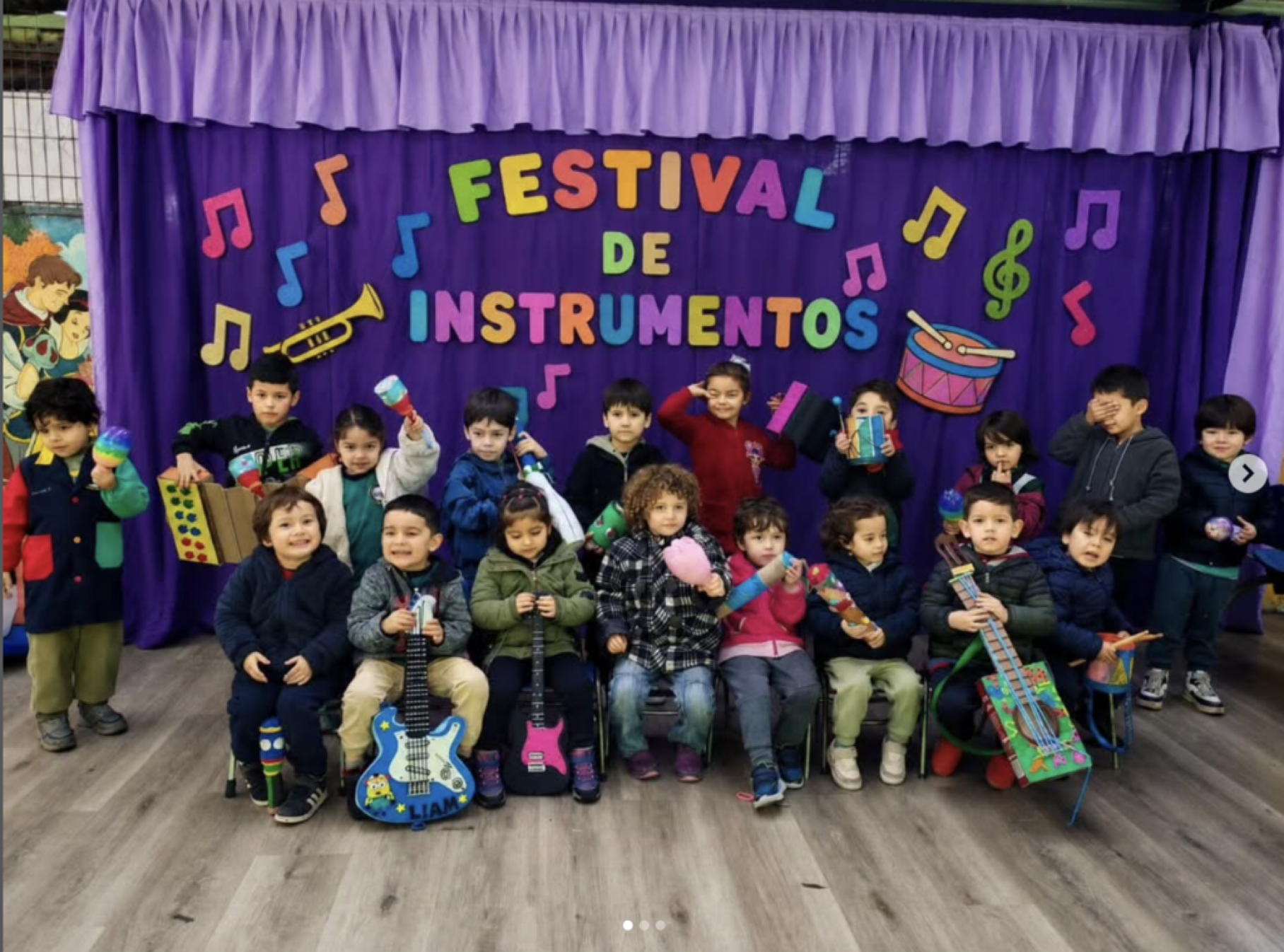 Grupo de niños en un escenario decorado con un telón morado, con letras de colores que dicen 'Festival de Instrumentos', algunos sosteniendo instrumentos musicales y otros con instrumentos de juguete, en celebración escolar.
