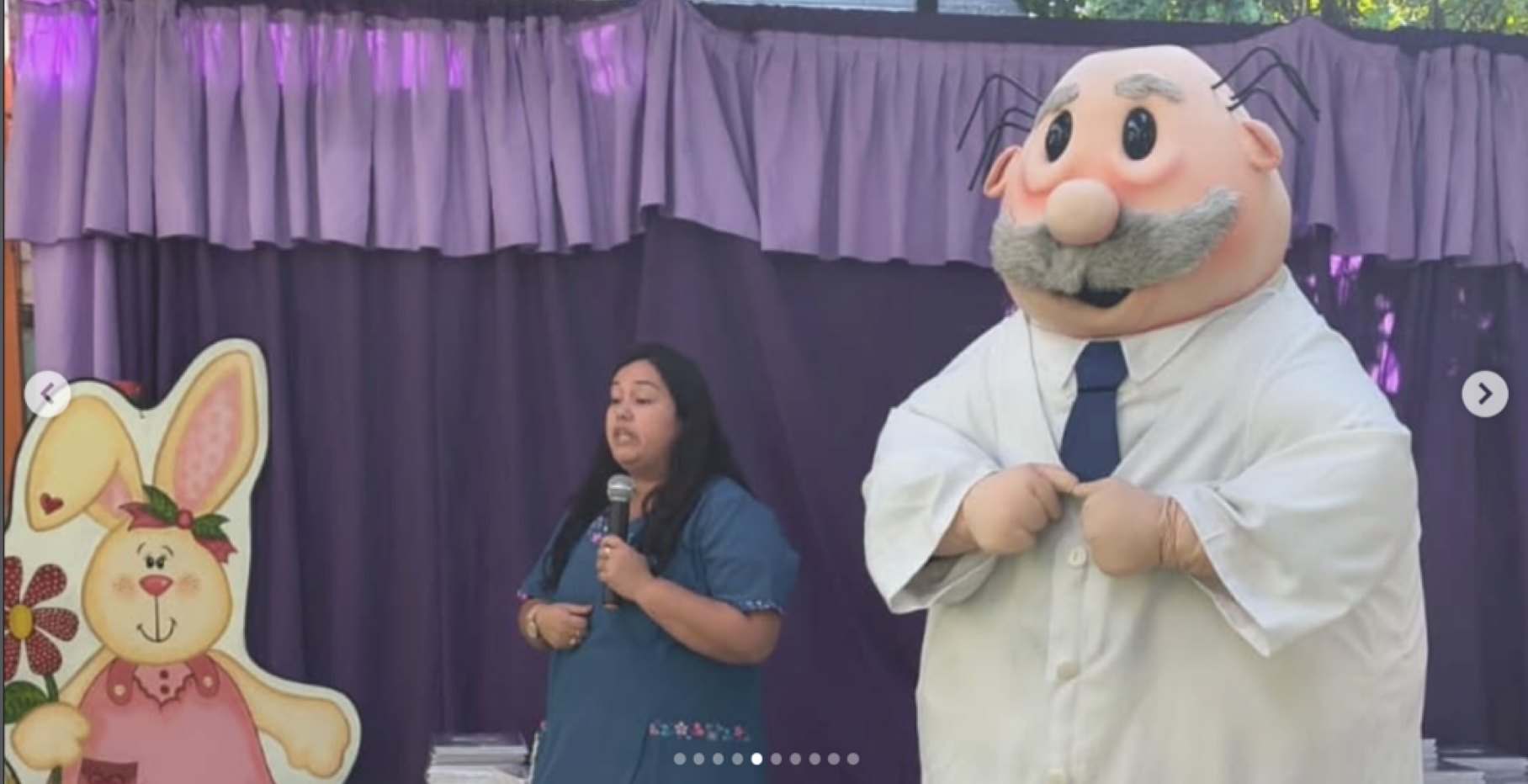 Una mujer hablando frente a un escenario con decoraciones de Pascua y un personaje disfrazado de abuelo con cabeza grande y bigote, con fondo de cortinas moradas.