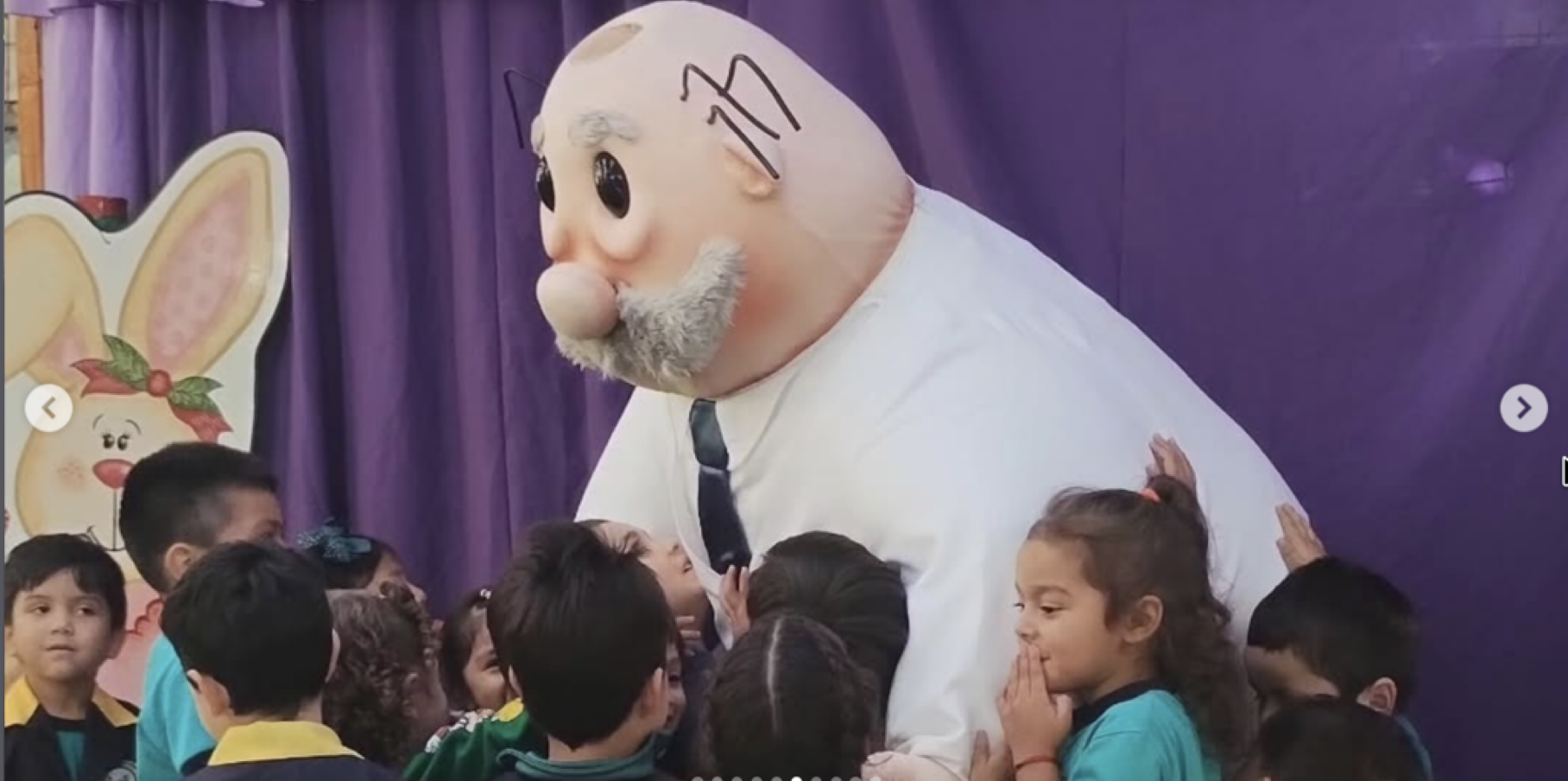 Un muñeco gigante con cara de abuelo, barba gris, vestido con camisa blanca y corbata, rodeado de niños en un evento navideño.