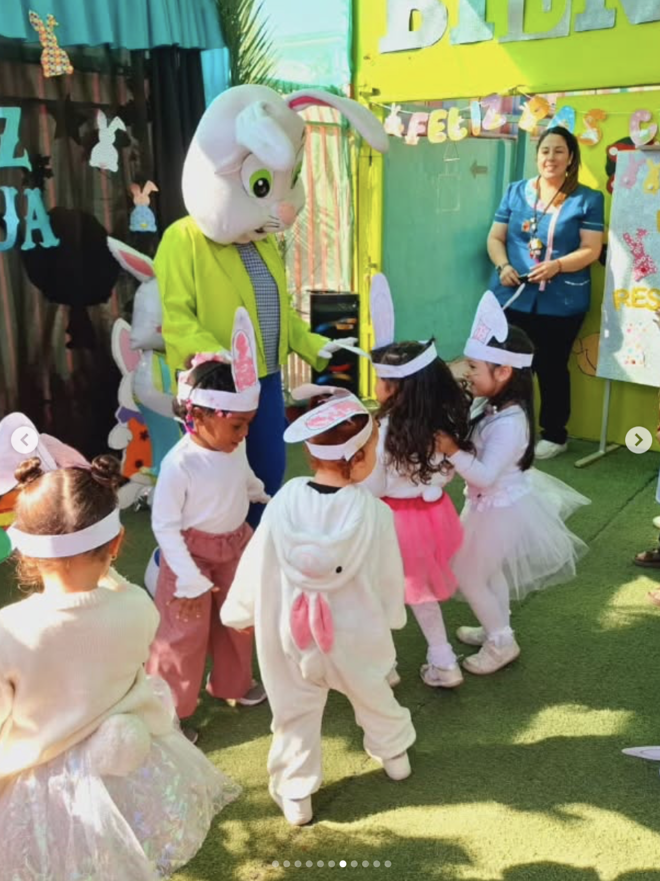 Niños disfrazados de conejitos y juegan en una celebración con decoraciones coloridas y un personaje disfrazado de conejo. Hay una mujer vestida con uniforme azul en el fondo, en un lugar decorado con banderines y dibujos de conejos.