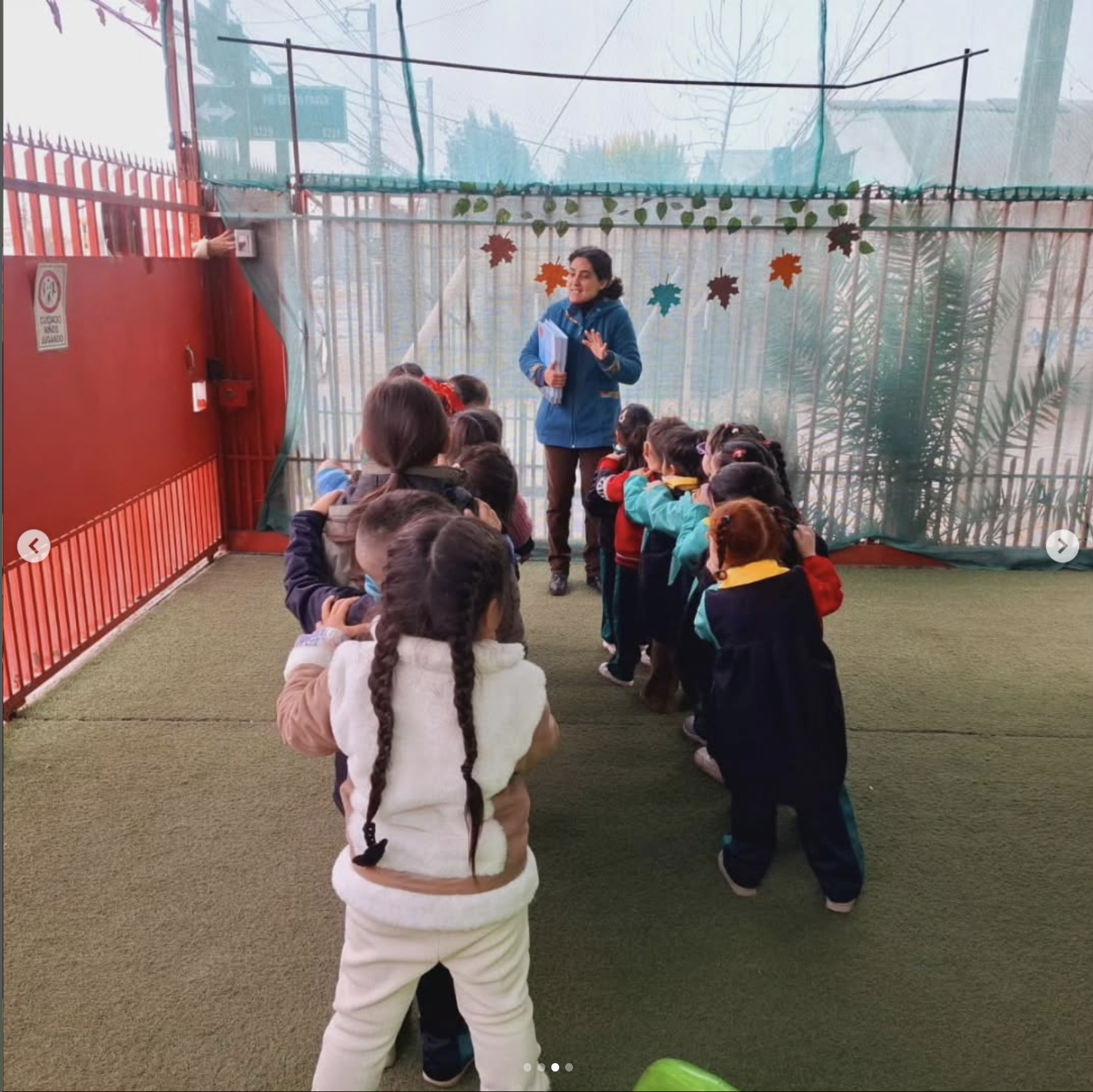 Niños en una fila formando una fila para escuchar a una mujer que les habla y les muestra un libro en una sala con decoración de hojas de colores de otoño colgadas del techo.