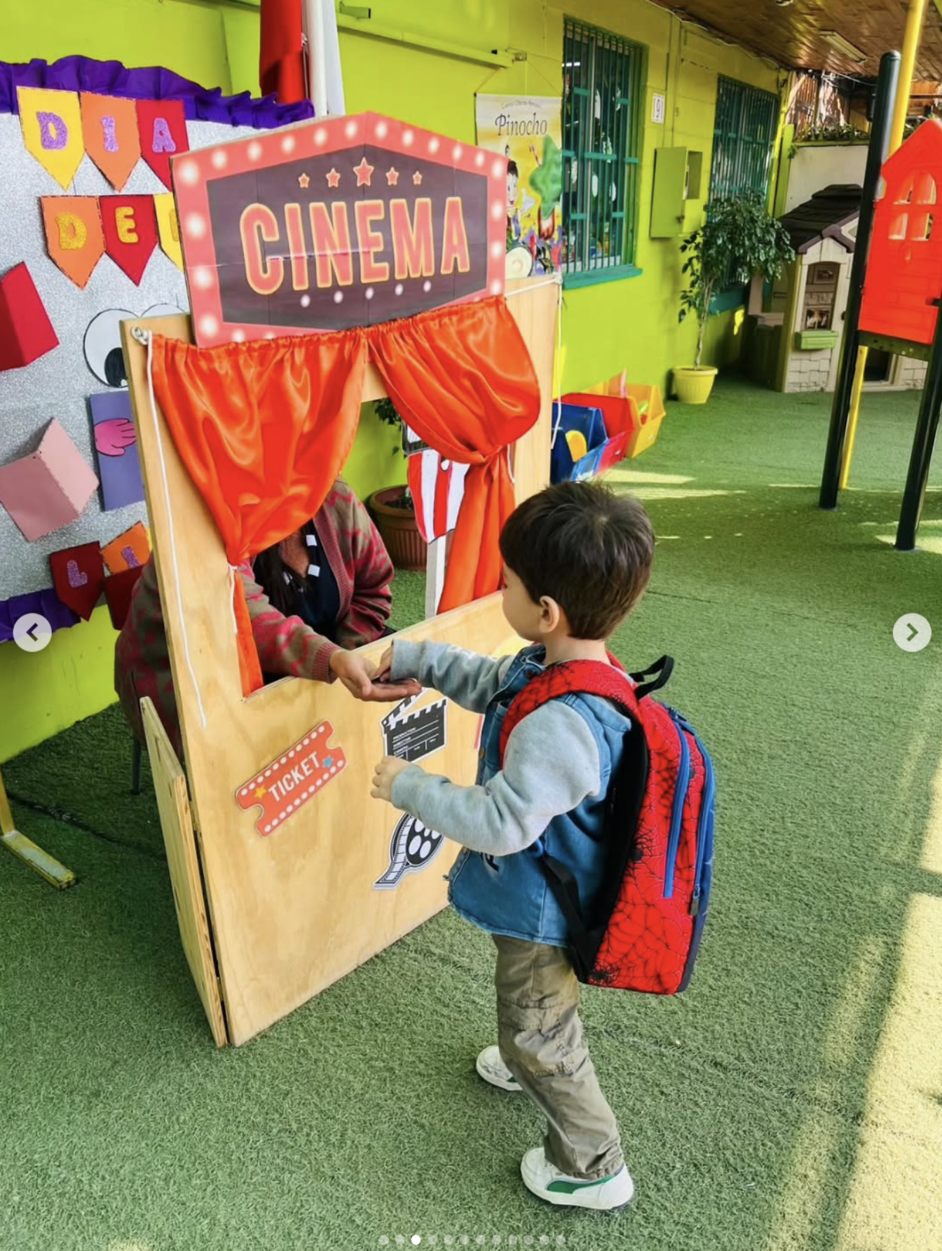 Un niño en un parque infantil jugando en una cabina de cine de juguetes con un cartel que dice 'CINEMA', con otros niños y decoraciones coloridas alrededor.