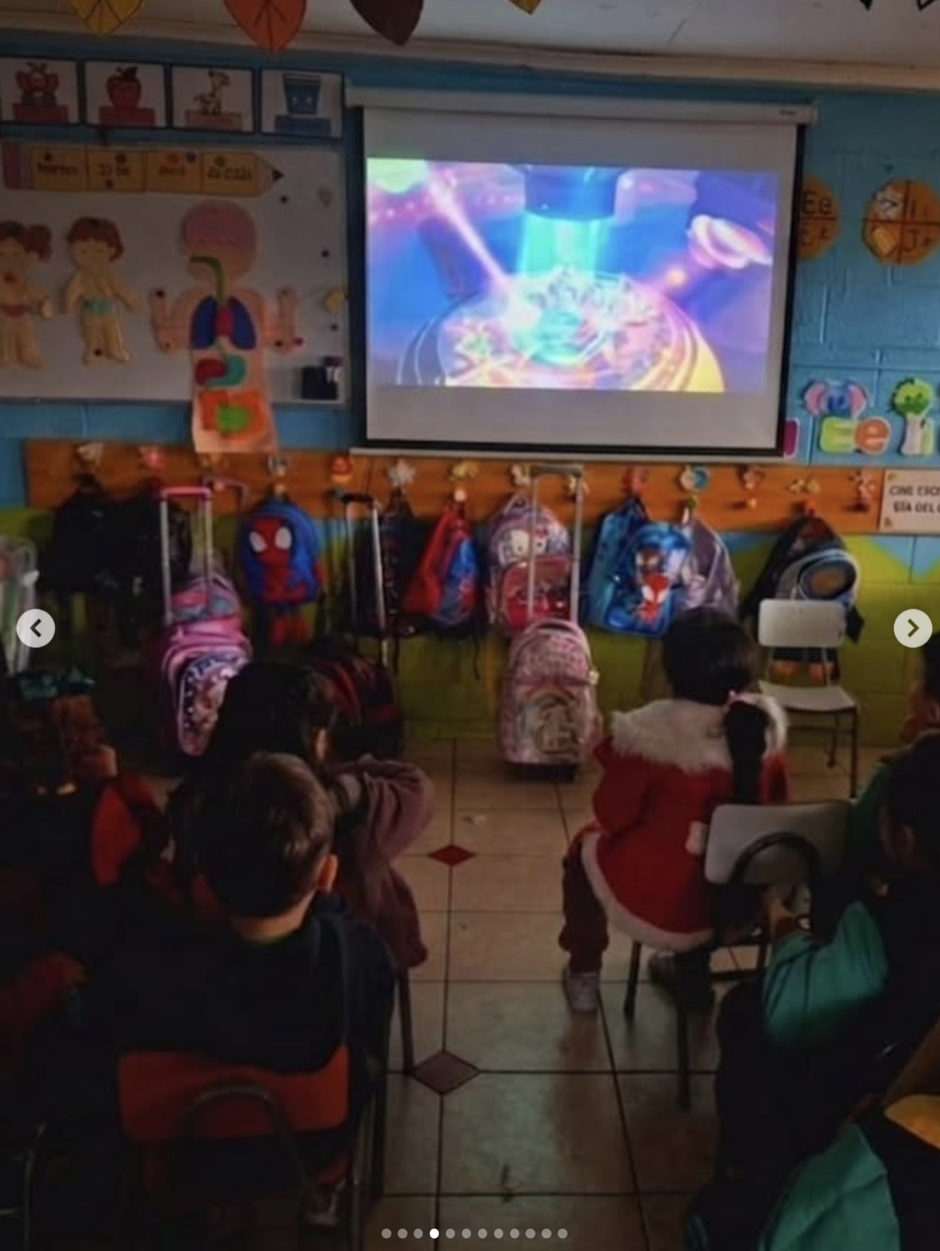 Niños en un aula viendo una presentación en una pantalla con un espectáculo de luces en colores vibrantes, con mochilas colgadas en un soporte y otros objetos decorativos en las paredes.