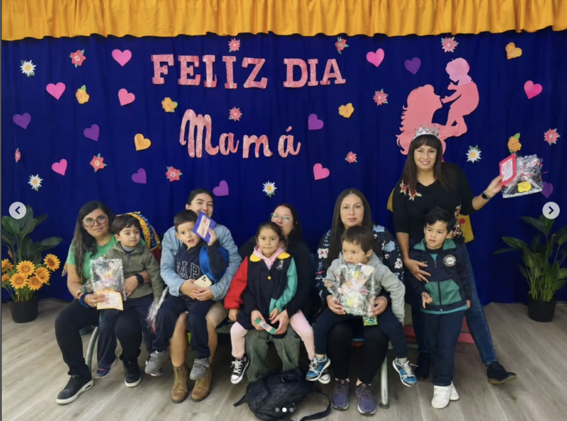 Grupo de niños y dos adultos en un evento con decoración de corazones, flores y un cartel que dice 'Feliz Día Mamá'; algunos niños sosteniendo regalos y premios.