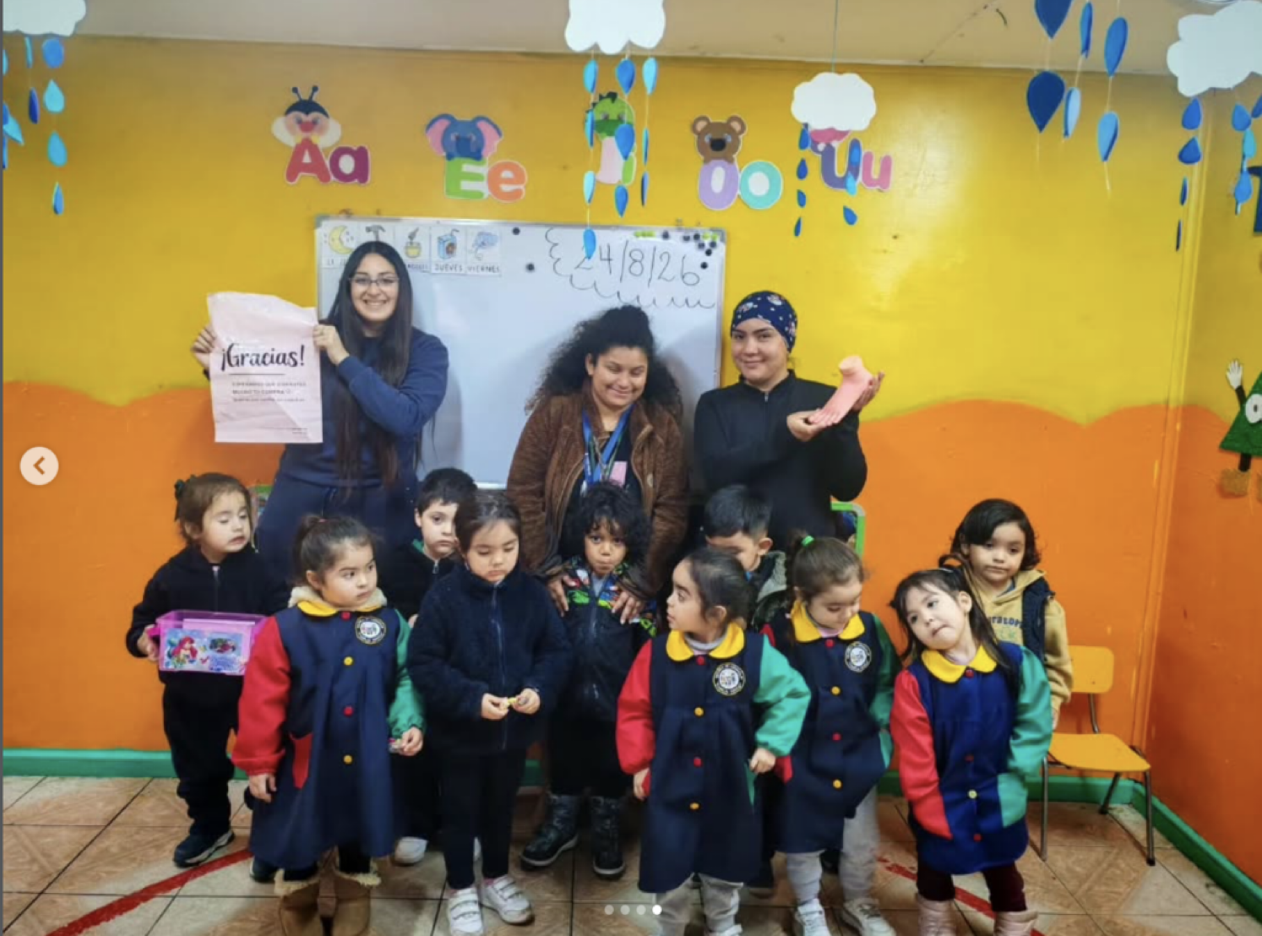 Grupo de niños en un salón de clases con decoraciones de nubes y letras del alfabeto, acompañados de adultos, algunos sosteniendo objetos y sonriendo.