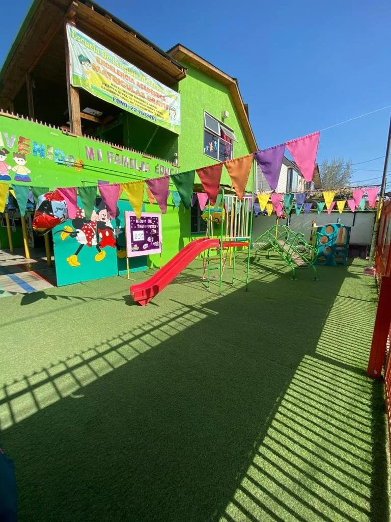 Área de juego infantil en un jardín con césped artificial, una estructura de escalada, resbaladilla roja y decoraciones de banderines coloridos, en frente de un edificio verde con cartel de una escuela.