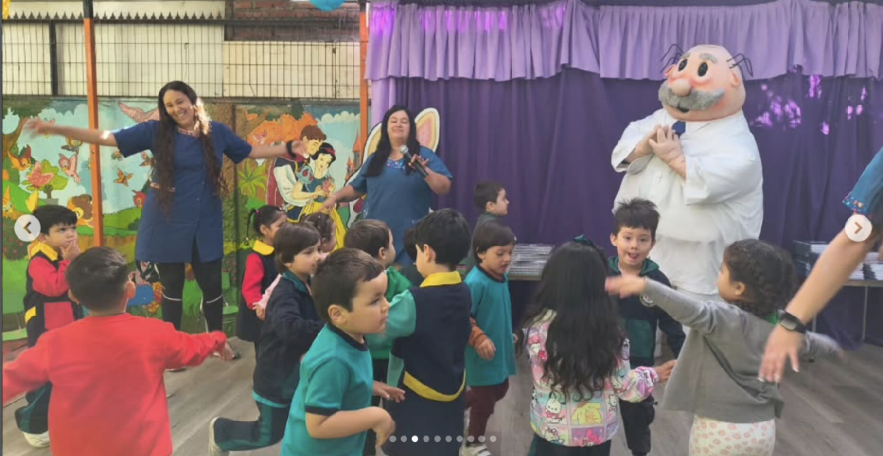 Niños y niñas en un salón de clases o teatro, participando en una actividad con personajes disfrazados, incluyendo un personaje con cabeza gigante de un anciano con barba y un vestido blanco, y una mujer con vestimenta azul que sostiene un micrófono.