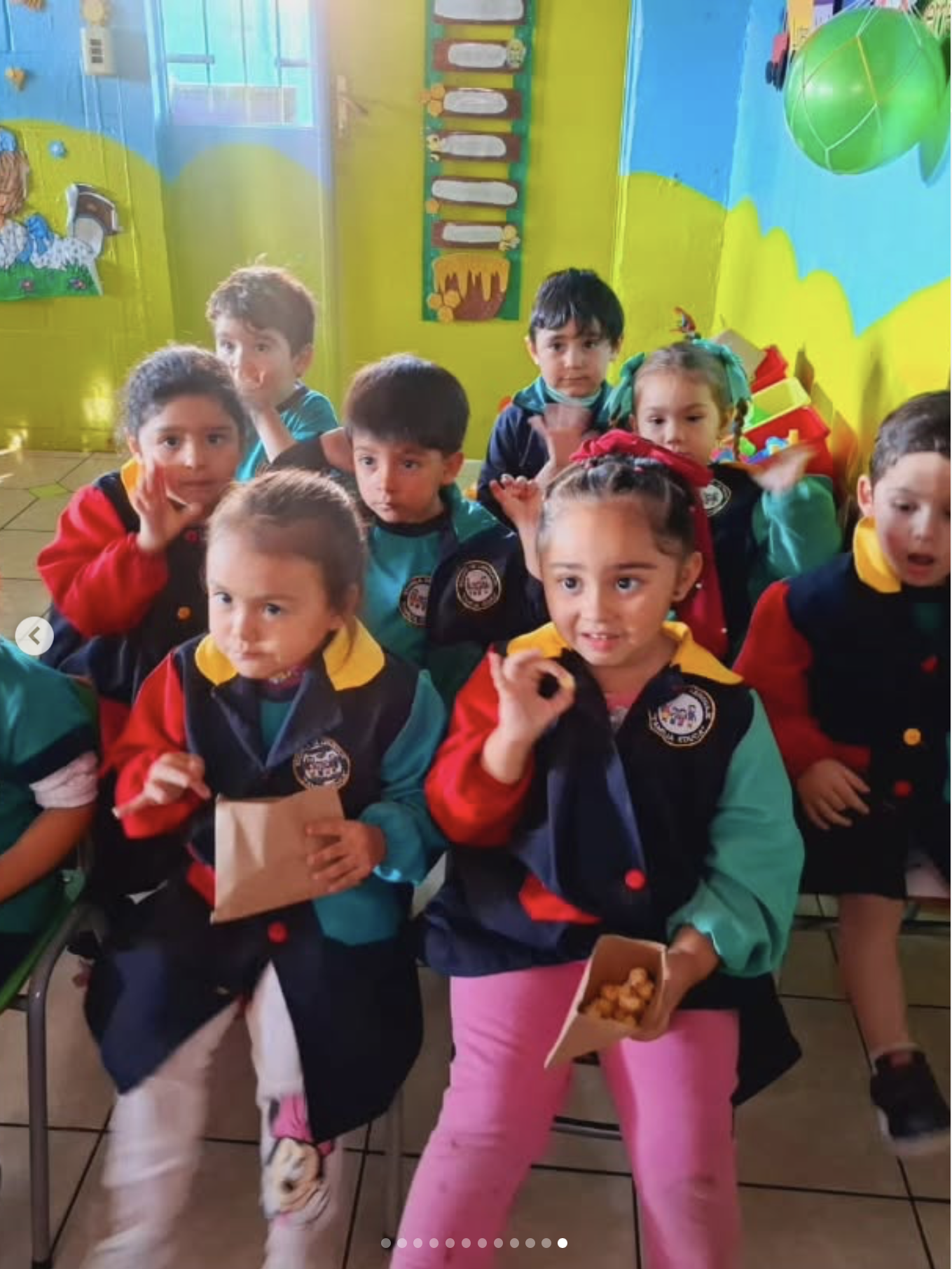 Niños en una aula de clases con paredes decoradas, algunos con disfraces de colores, sosteniendo bolsas y comida, algunos hacen gestos hacia la cámara.