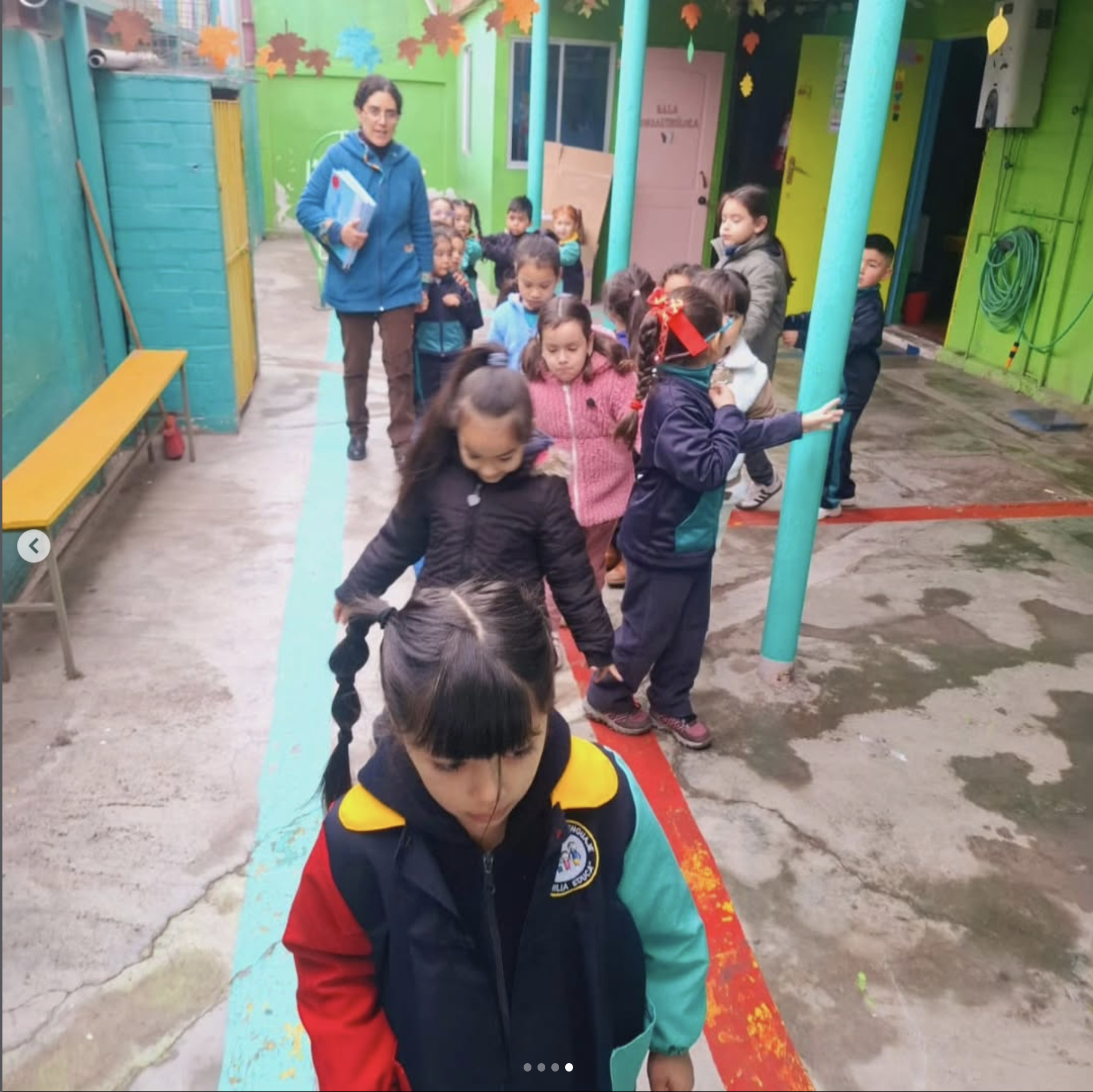 Niños en fila en un patio escolar, algunos con máscaras, acompañados por adultos en un ambiente colorido y alegre, con paredes verdes y decoraciones de hojas en la pared.