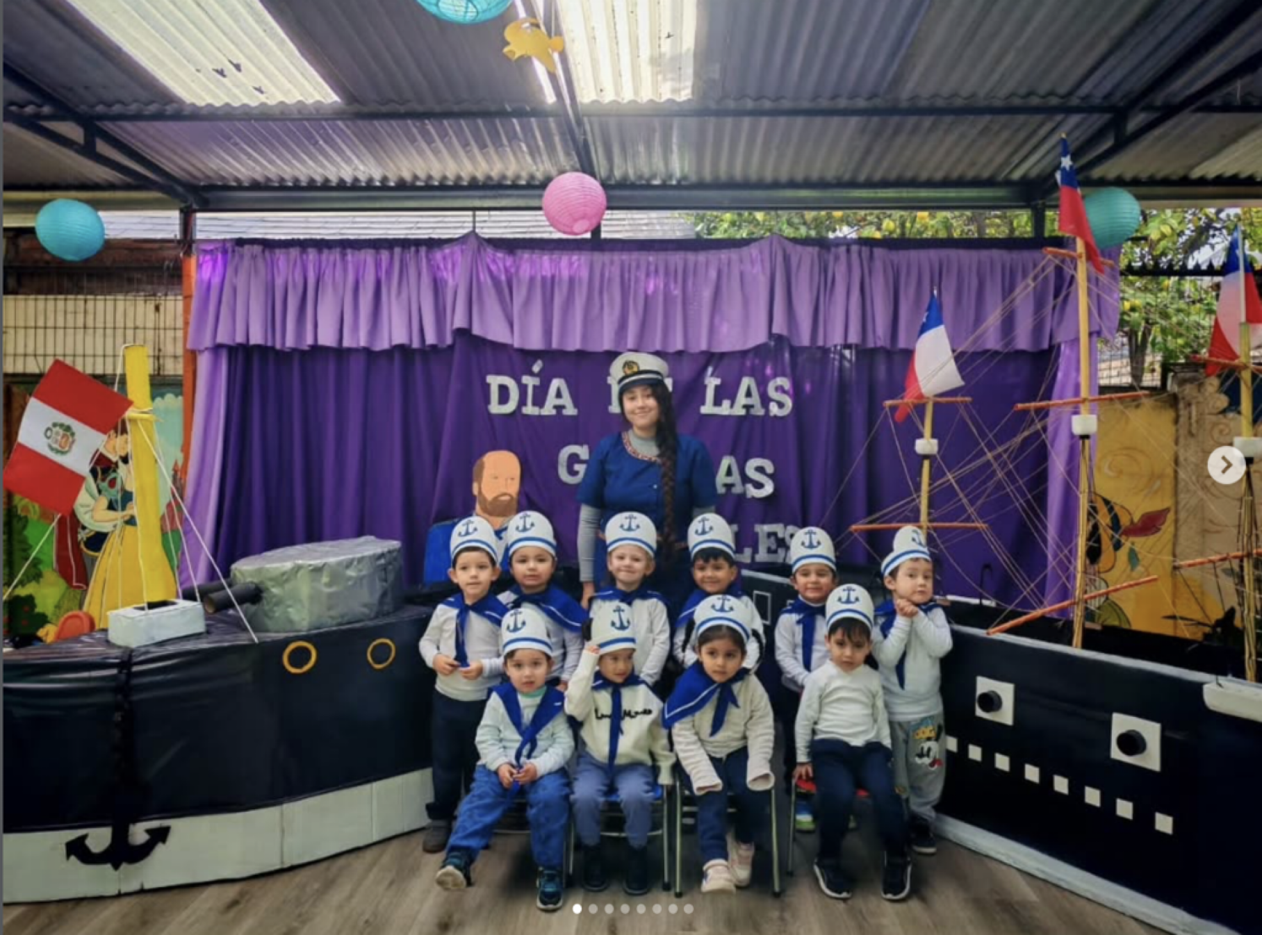 Grupo de niños y una mujer vestida con uniforme de capitán de barco en una celebración en honor al día de las guerras navales, con decoraciones de temática marina y banderas en el fondo.