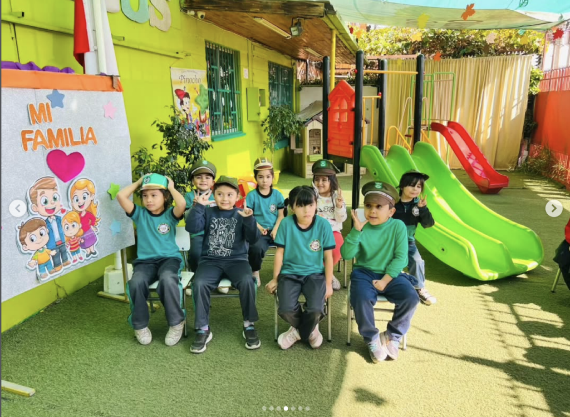 Grupo de niños en un patio de escuela con estructura de juegos, algunos usan gorra. Pared verde con cartel de 'Mi Familia' y personajes de dibujos, y un mural de Pinocho. Vegetación y una cerca en el fondo.