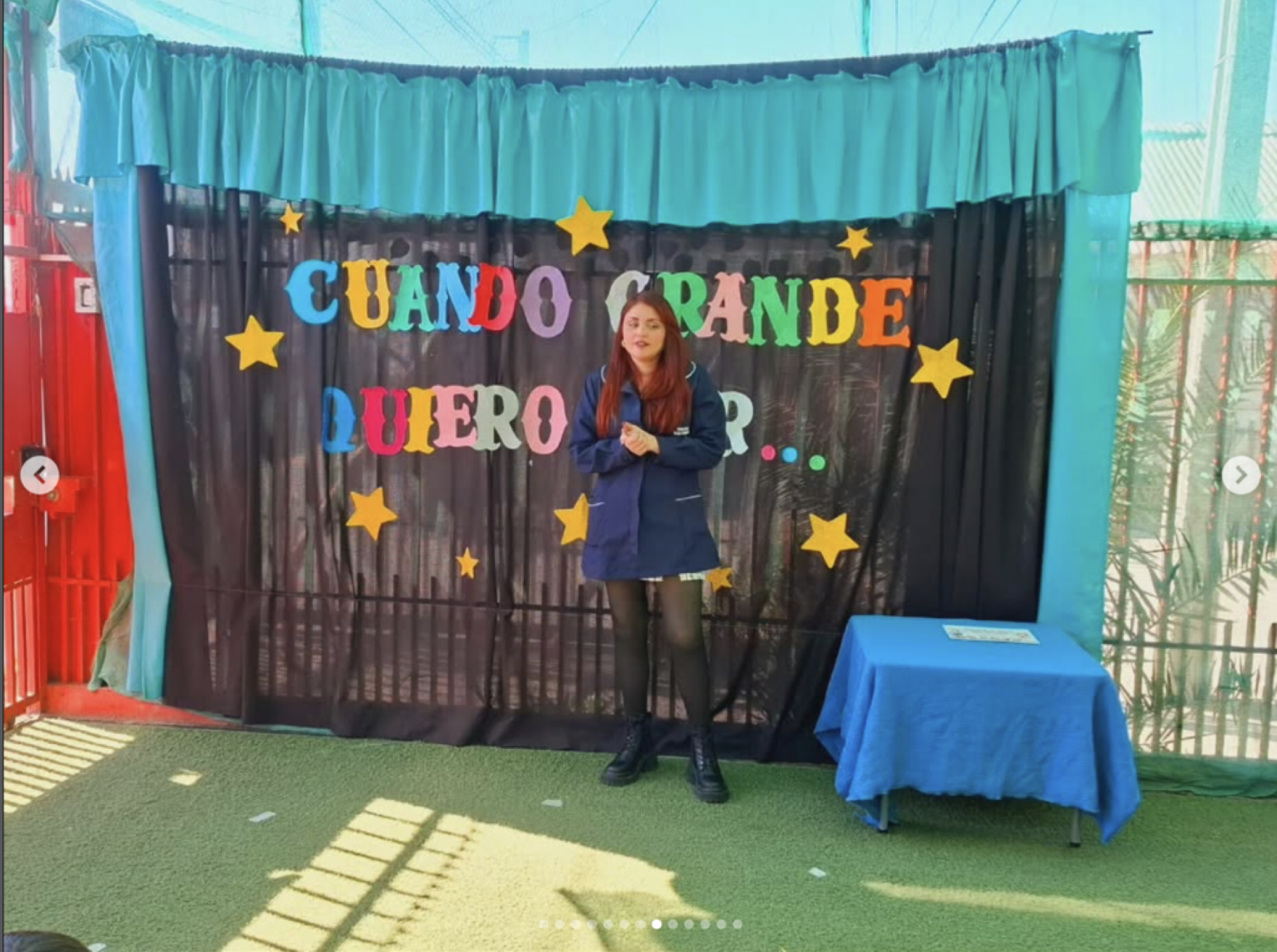 Escenario con un fondo de cortina negra y decoraciones de estrellas amarillas, con un cartel colorido que dice "Cuando grande quiero ser...", una mujer con cabello largo y vestida con abrigo azul y falda negra, de pie frente al escenario, en un espac