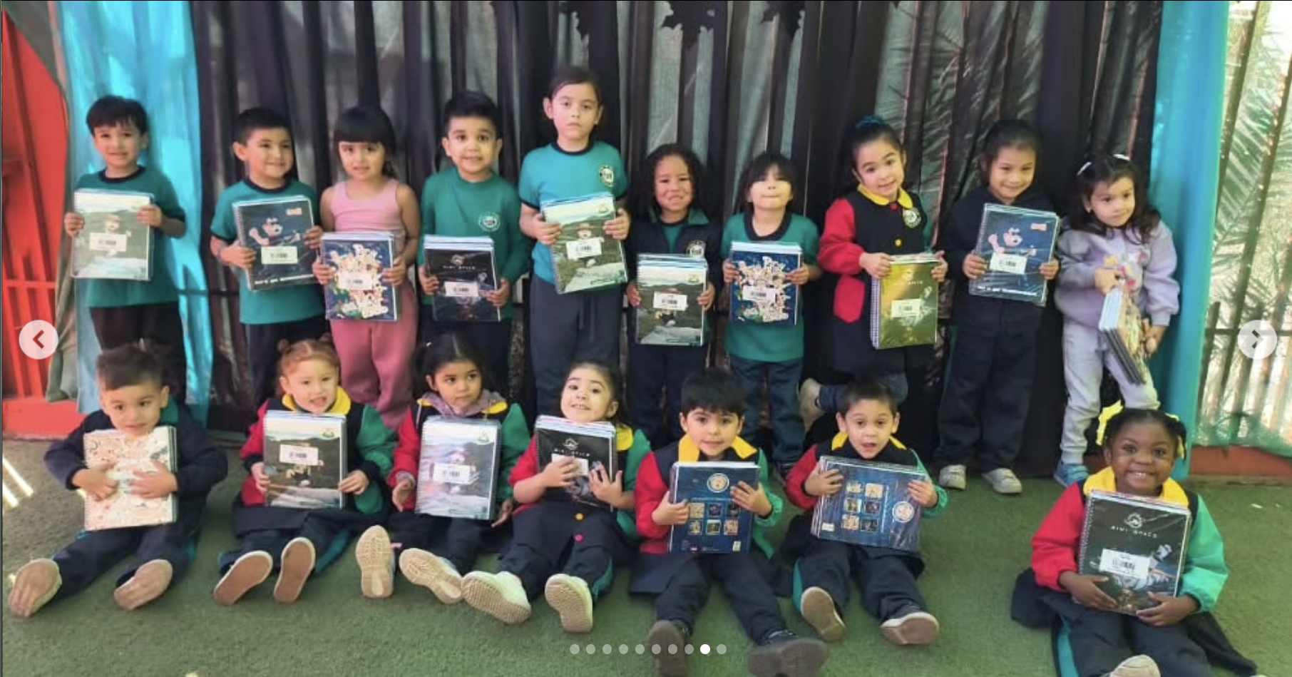Grupo de niños y niñas en una escuela, algunos sentados en el suelo y otros de pie, sosteniendo libros y cuadernos, con fondo de cortinas y paredes de colores.