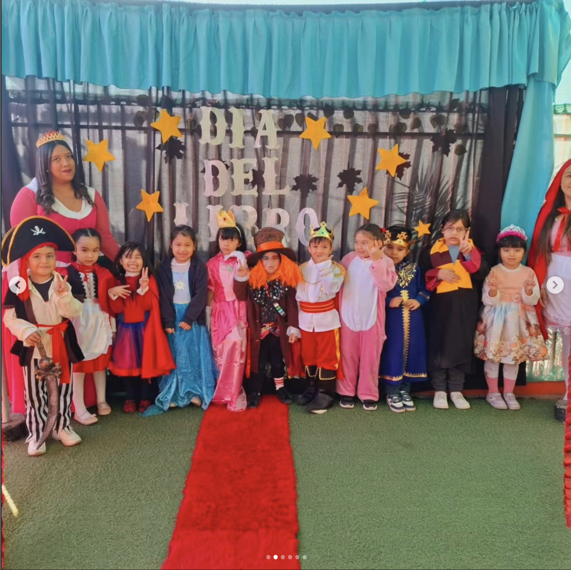 Niños y adultos disfrazados en una fiesta del día del niño con decoraciones de estrellas y letras, en un escenario con cortinas azules y fondo decorado con letras gigantes que dicen 'Día del Niño'.