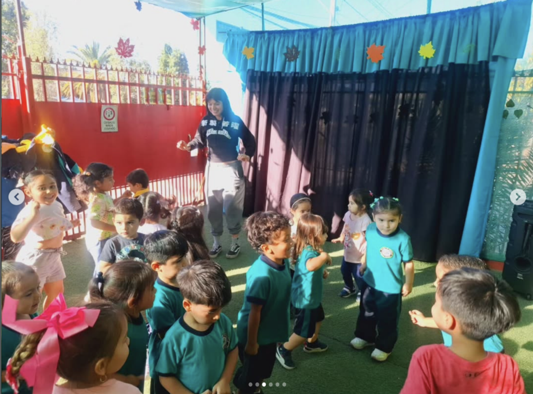 Niños y una mujer en un área de juegos cubierta, decorada con hojas de colores, participando en una actividad escolar al aire libre.