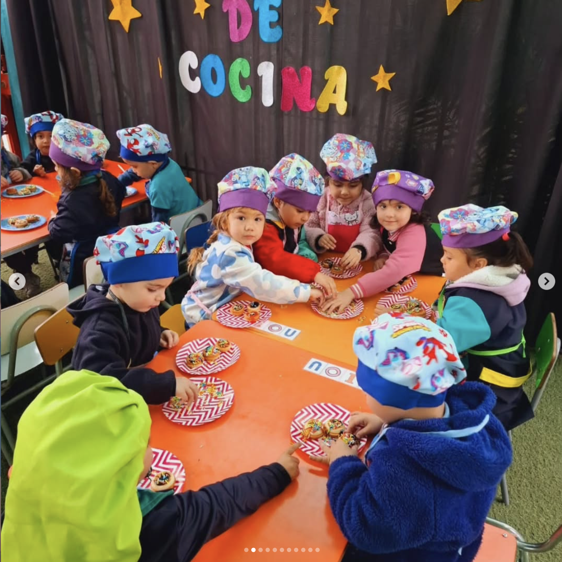 Niños en una fiesta infantil decorada con letras de colores que dicen 'DÍA DE COLORES', usando gorros de papel con diseño colorido, comiendo galletas decoradas en un mesa con mantel de rayas rojas y blancas.