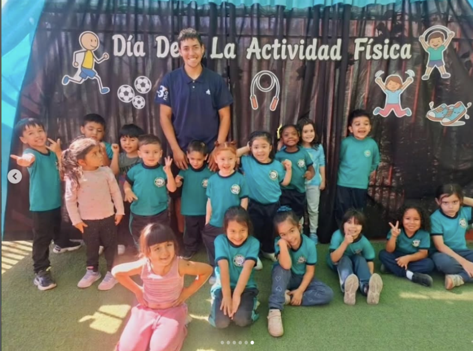 Niños y una adulta en celebración del Día de la Actividad Física, algunos niños con camisetas azules y otros con ropa casual, en un espacio con decoraciones que incluyen dibujos de niños jugando y elementos deportivos.