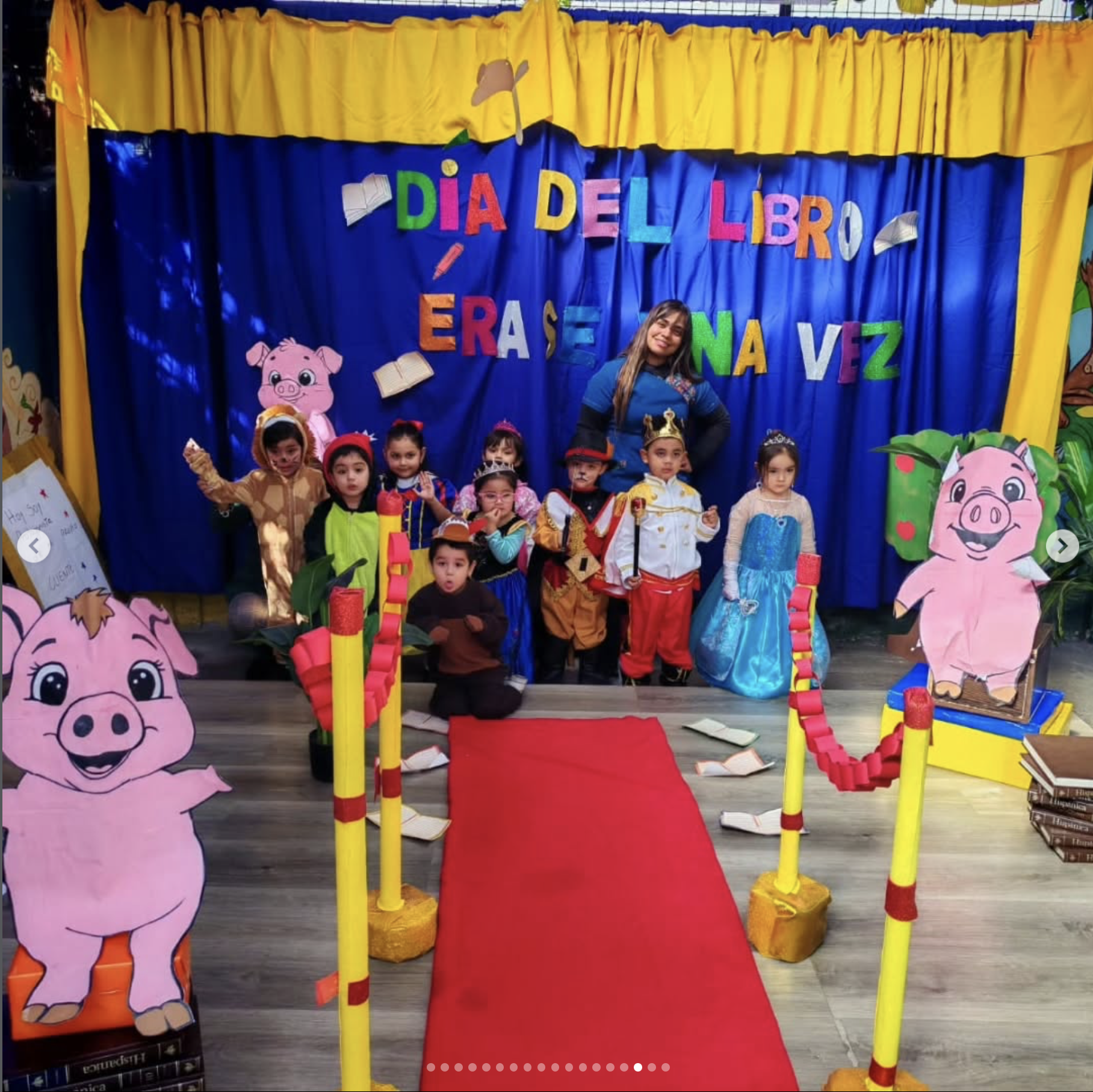 Celebração de Día del Libro con niños disfrazados de personajes de cuentos, decoraciones coloridas, telón de fondo azul con letras multicolores que dicen 'Día del Libro'.