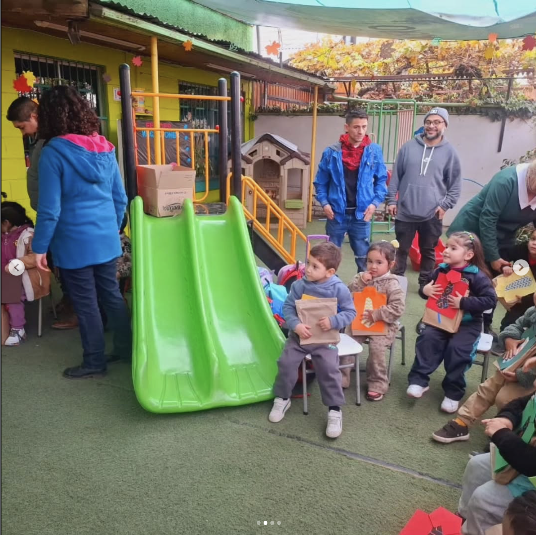 Niños y adultos en un área de juegos y socialización infantil con resbaladilla verde, vegetación de otoño y paredes verdes y grises.
