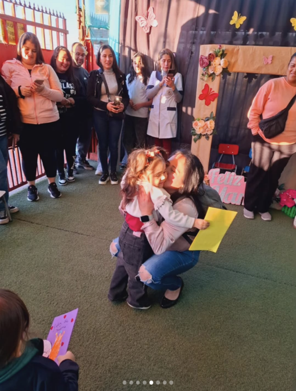 Una mujer y una niña se abrazan en una celebración del Día de las Madres, rodeadas de varias personas que observan y toman fotos, en un lugar decorado con flores y mariposas, y una señal que dice "Feliz Día".