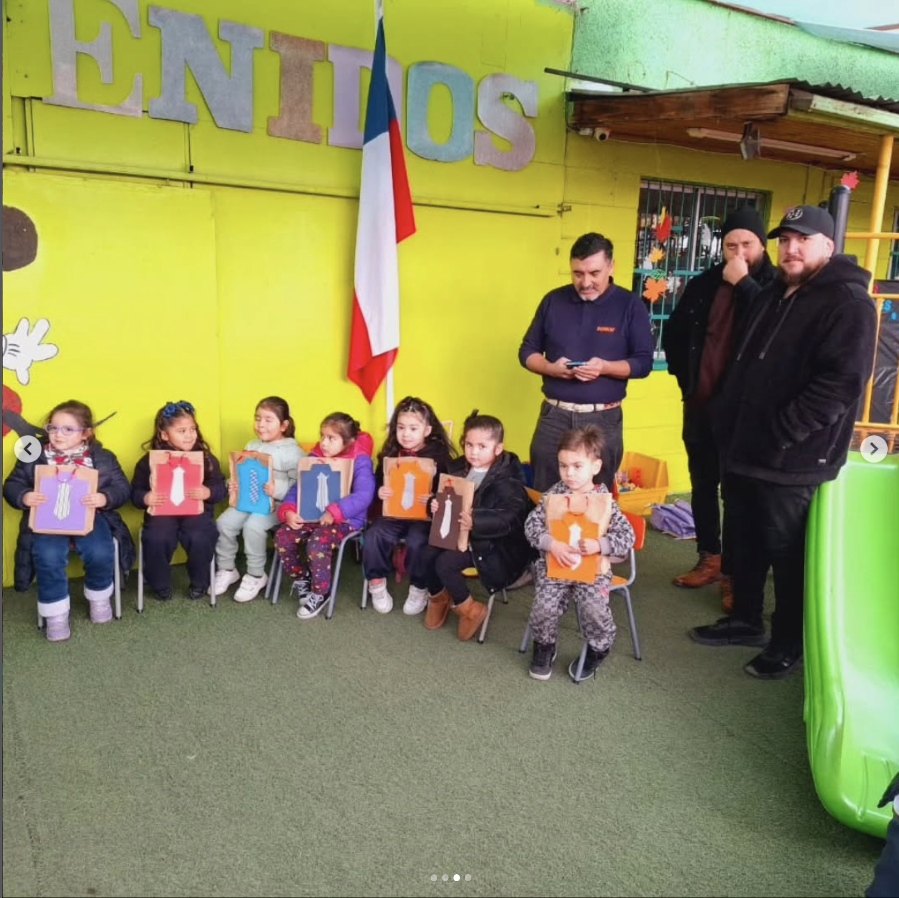 Grupo de niños y adultos en una clase o evento escolar con decoración colorida y bandera, sosteniendo cuadros con símbolos y colores, en un salón con paredes verdes y piso de césped artificial.