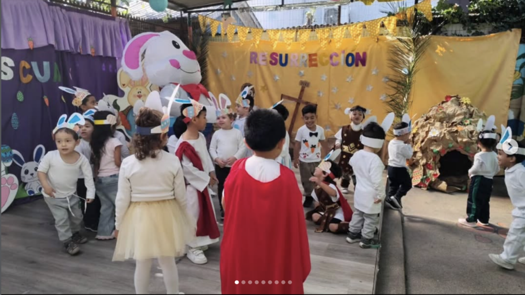 Niños en una celebración con decoraciones de Pascua, disfrazados de conejos y otros personajes, en un escenario con fondo amarillo y elementos de cabaña y árboles.