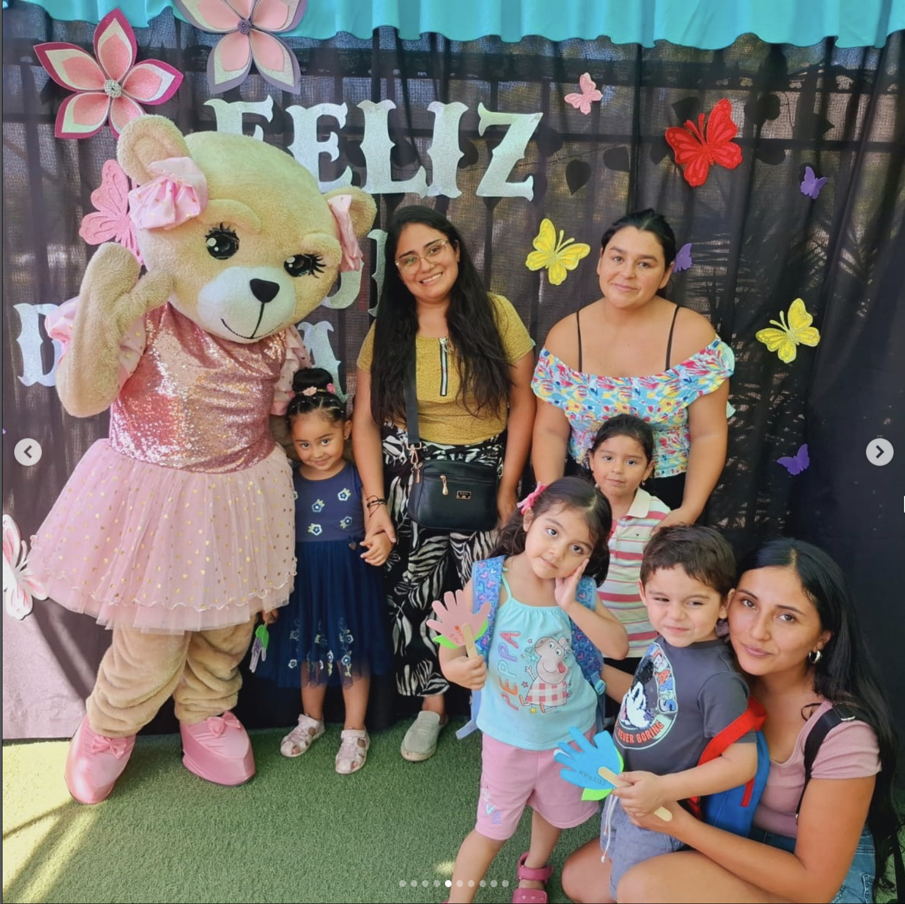 Grupo de niños y adultos en una celebración con decoraciones coloridas de mariposas y letras que dicen 'FELIZ'. Una persona con disfraz de osito de peluche rosa y amarillo, con vestido rosa, posa para la fotografía.