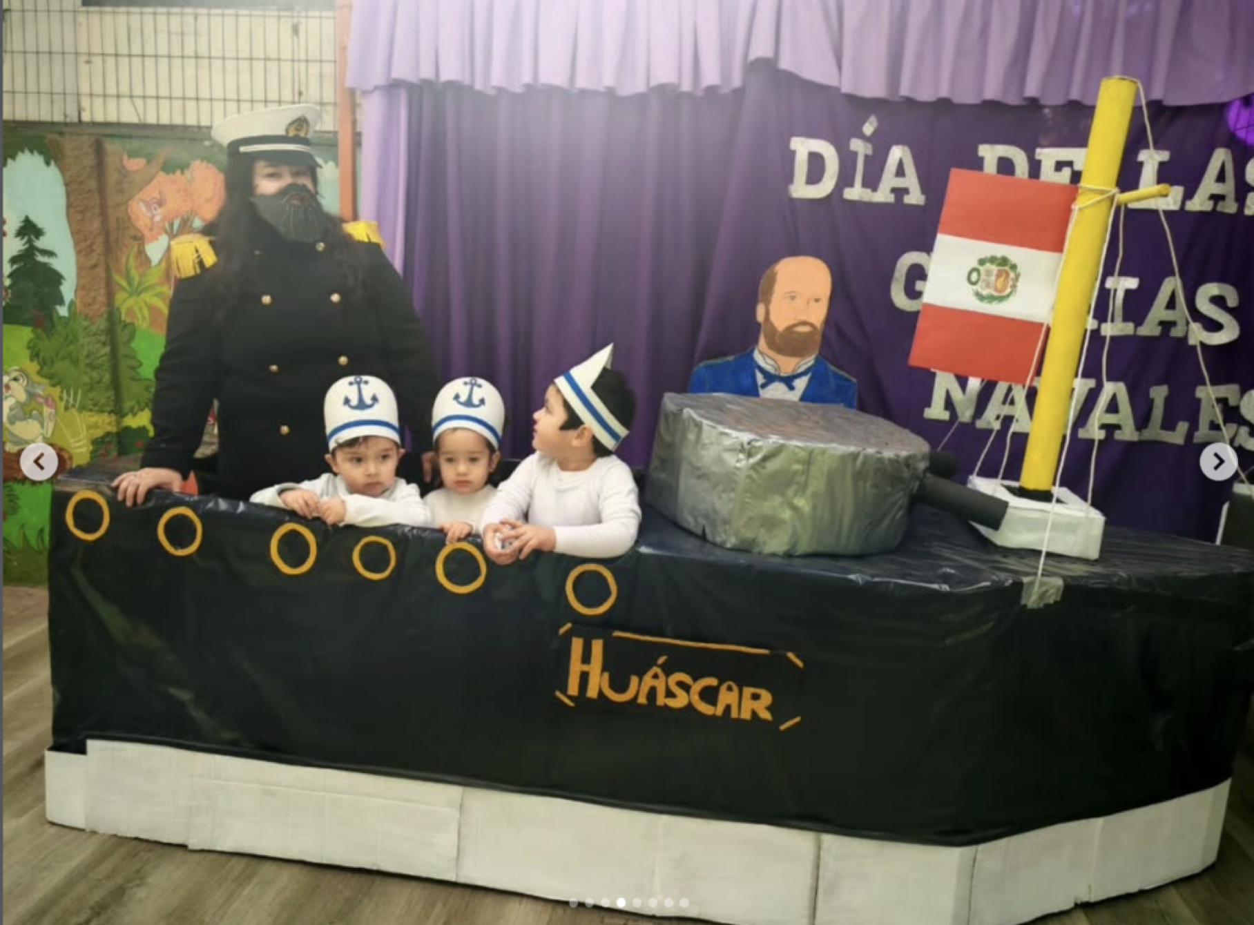 Niños en una mesa decorada con temática náutica, vestida con camisetas y gorras con ancla, participando en una celebración del día de las velas, con un adulto vestido de marinero frente a un telón morado con letras en español y un banderín peruano.