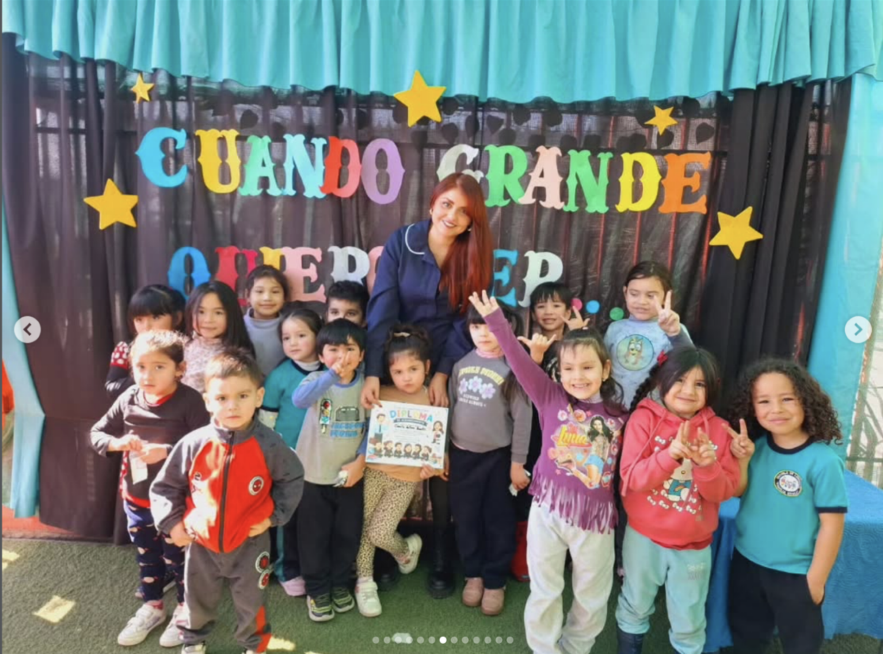 Grupo de niños con una maestra en una celebración escolar, posando frente a un fondo con letras de colores que dicen 'CUANDO GRANDE QUIERO SER'.