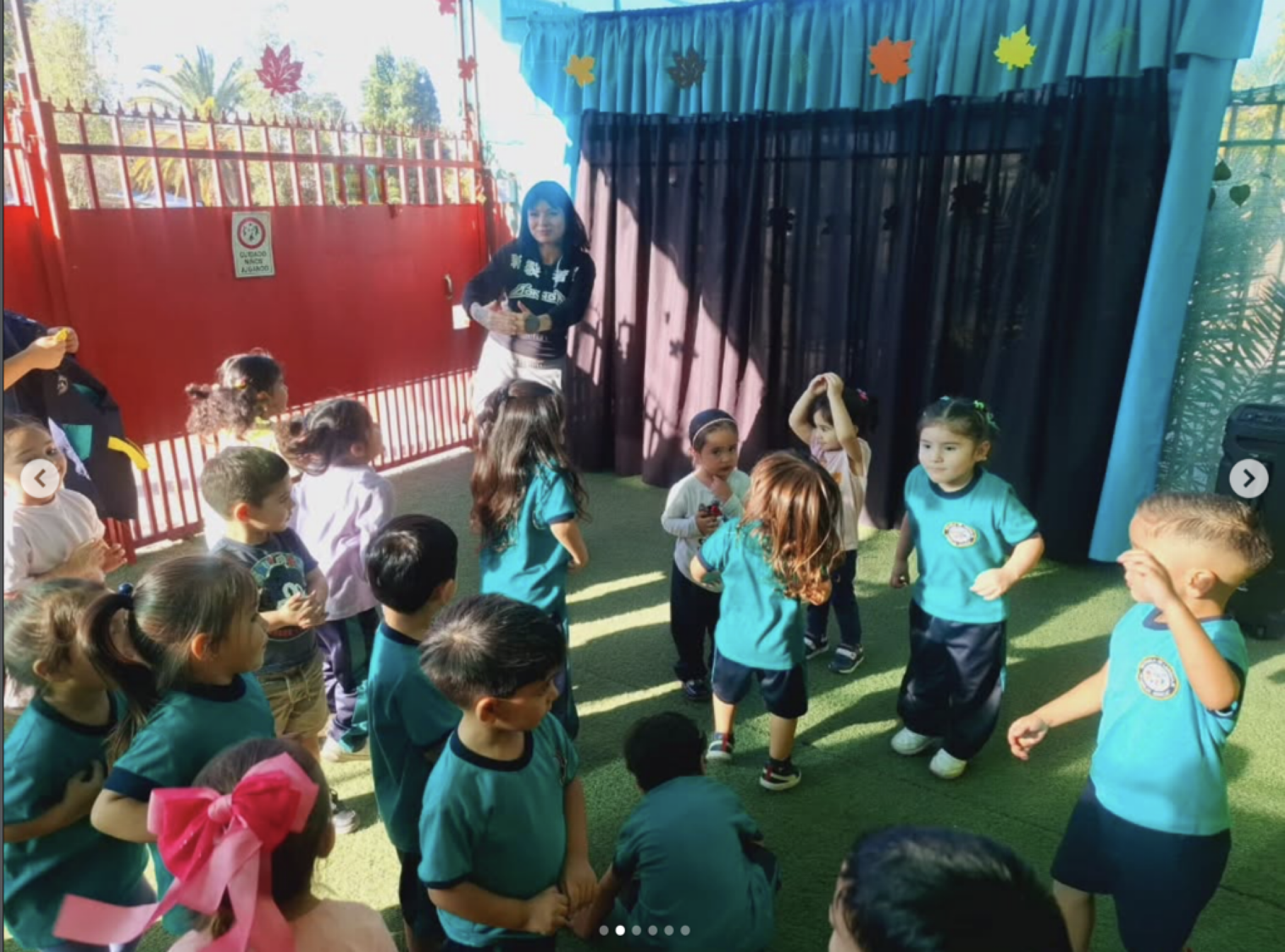 Grupo de niños en un área de juego cubierta, algunos con uniformes escolares azules y otros con ropa casual, atendiendo a una mujer que parece ser su profesora o cuidadora, en un ambiente colorido con decoraciones de hojas en el techo.