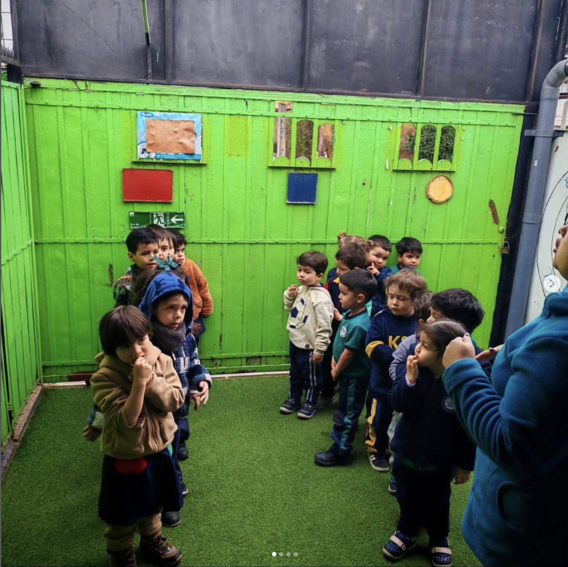 Niños congregados en un espacio cerrado con paredes verdes y piso de césped artificial, algunos están de pie, algunos en grupos, probablemente en una actividad escolar o recreativa.