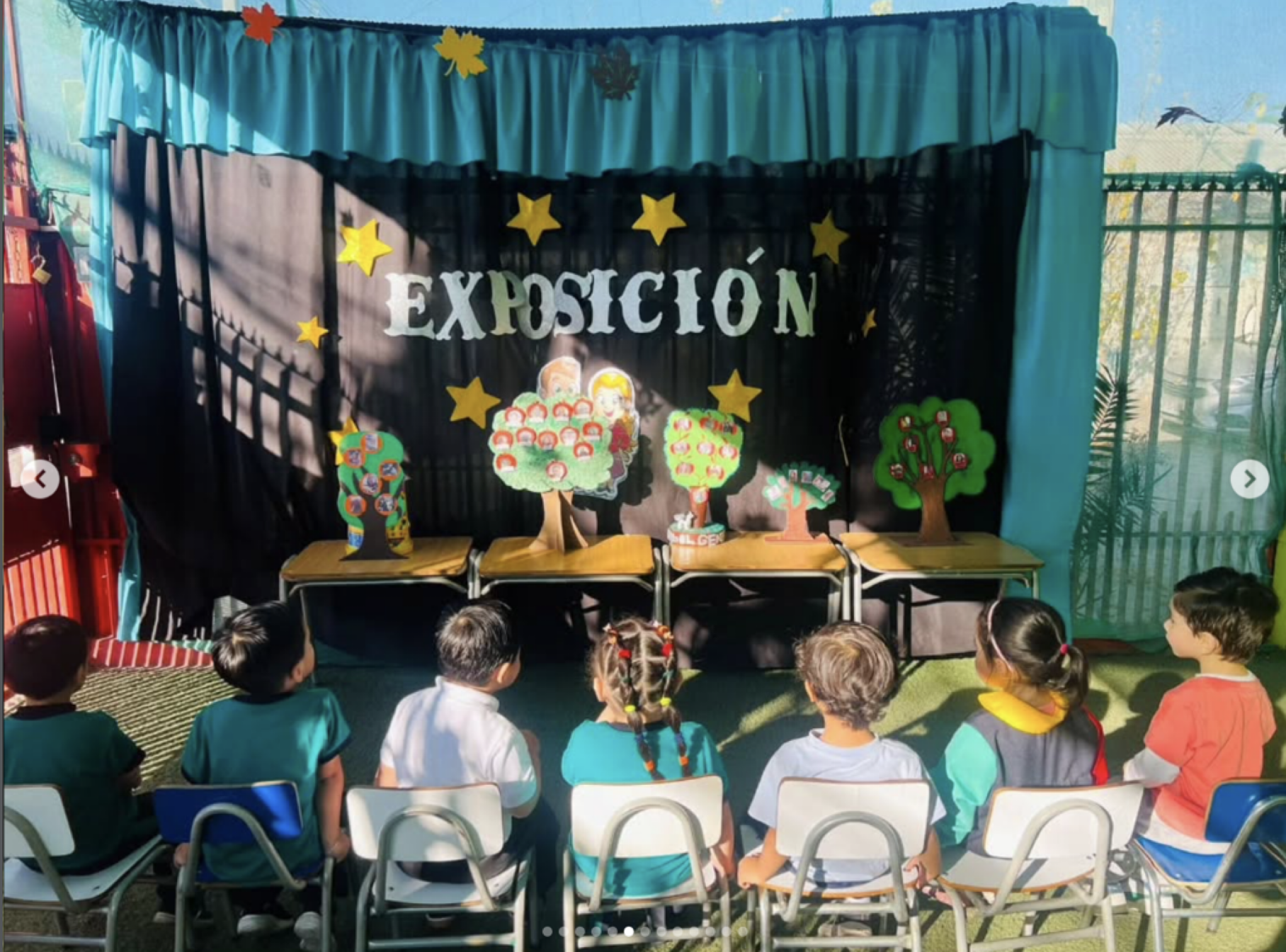 Niños en una escuela viendo una obra de teatro con decoraciones de árboles y estrellas, y un cartel que dice 'Exposición' en el fondo.