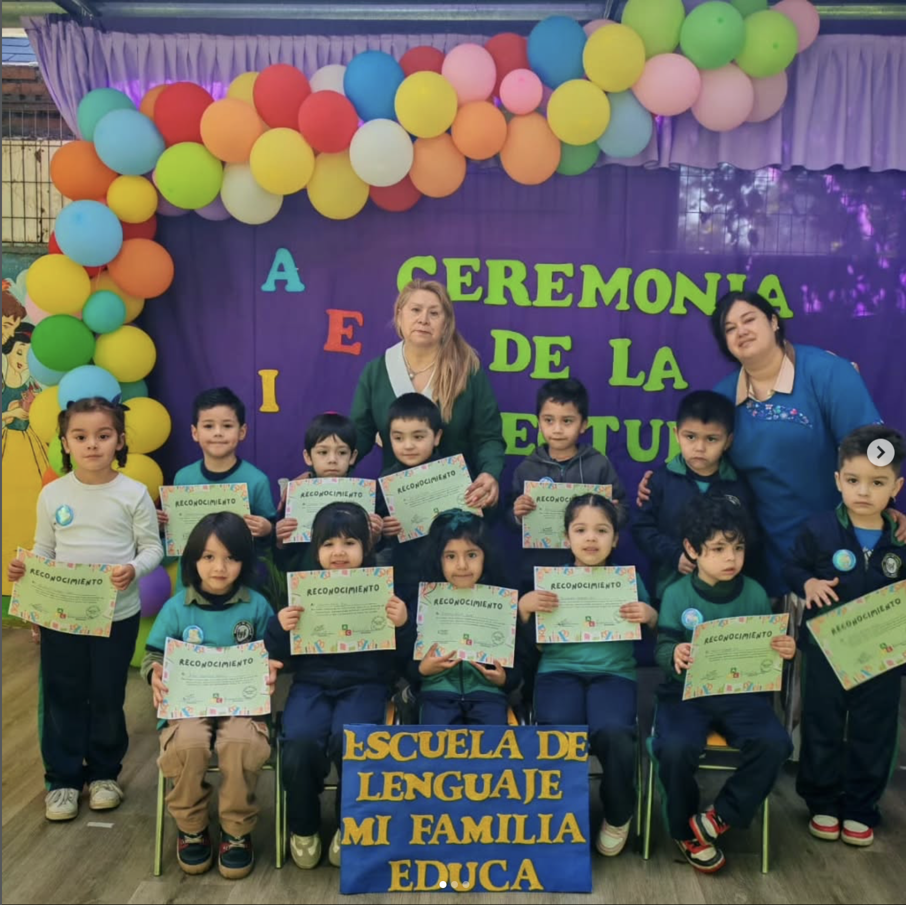 Niños y adultos en una ceremonia con decoración de globos de colores en un fondo púrpura que dice 'Ceremonia de la escuela de lengua, mi familia, educa'. Los niños sostienen certificados de reconocimiento.