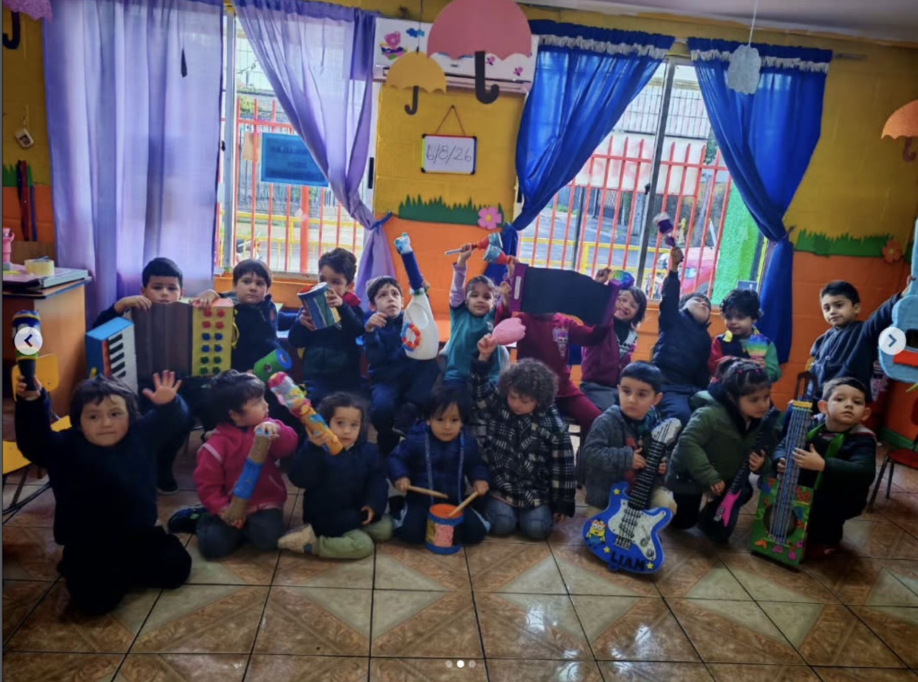 Grupo de niños en una clase de preescolar, algunos sosteniendo instrumentos musicales y juguetes, decoraciones coloridas y cortinas azules en el fondo, en un ambiente alegre y educativo.