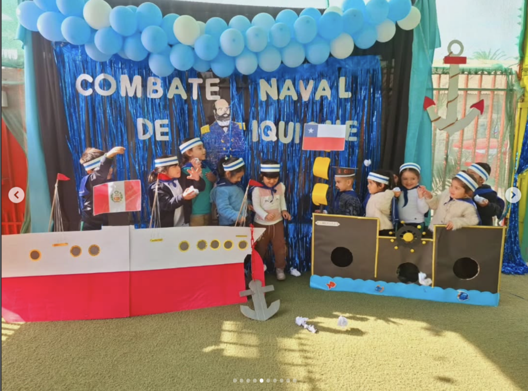 Niños en un escenario decorado con temática naval, con globos azules, un telón con guirnaldas azules, y figuras de barcos y anclas, participando en una actividad escolar sobre la marina y la naval.