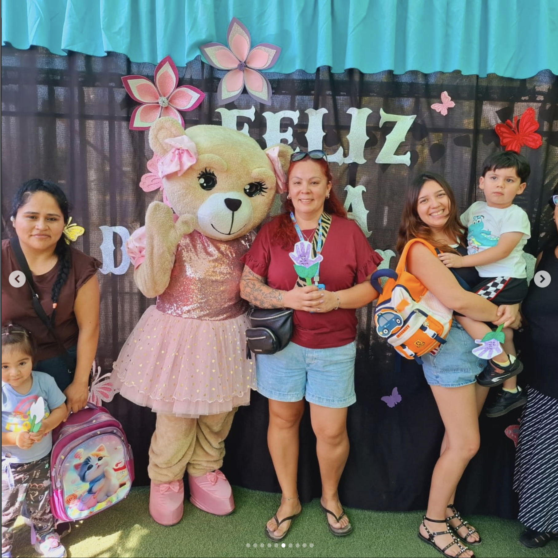 Grupo de personas con una mascota de personaje de peluche en un escenario decorado con flores y letras, en una celebración infantil.