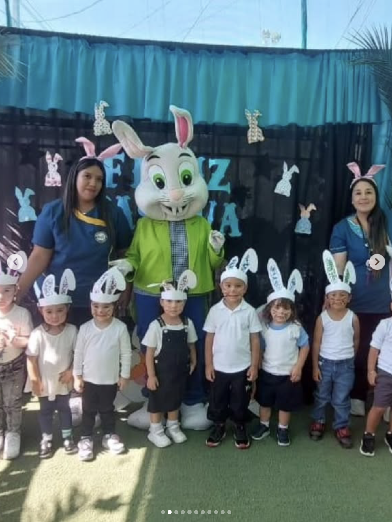 Grupo de niños y adultos en celebración de Pascua con decoraciones de conejitos y orejas de conejo en un fondo decorativo colorido.