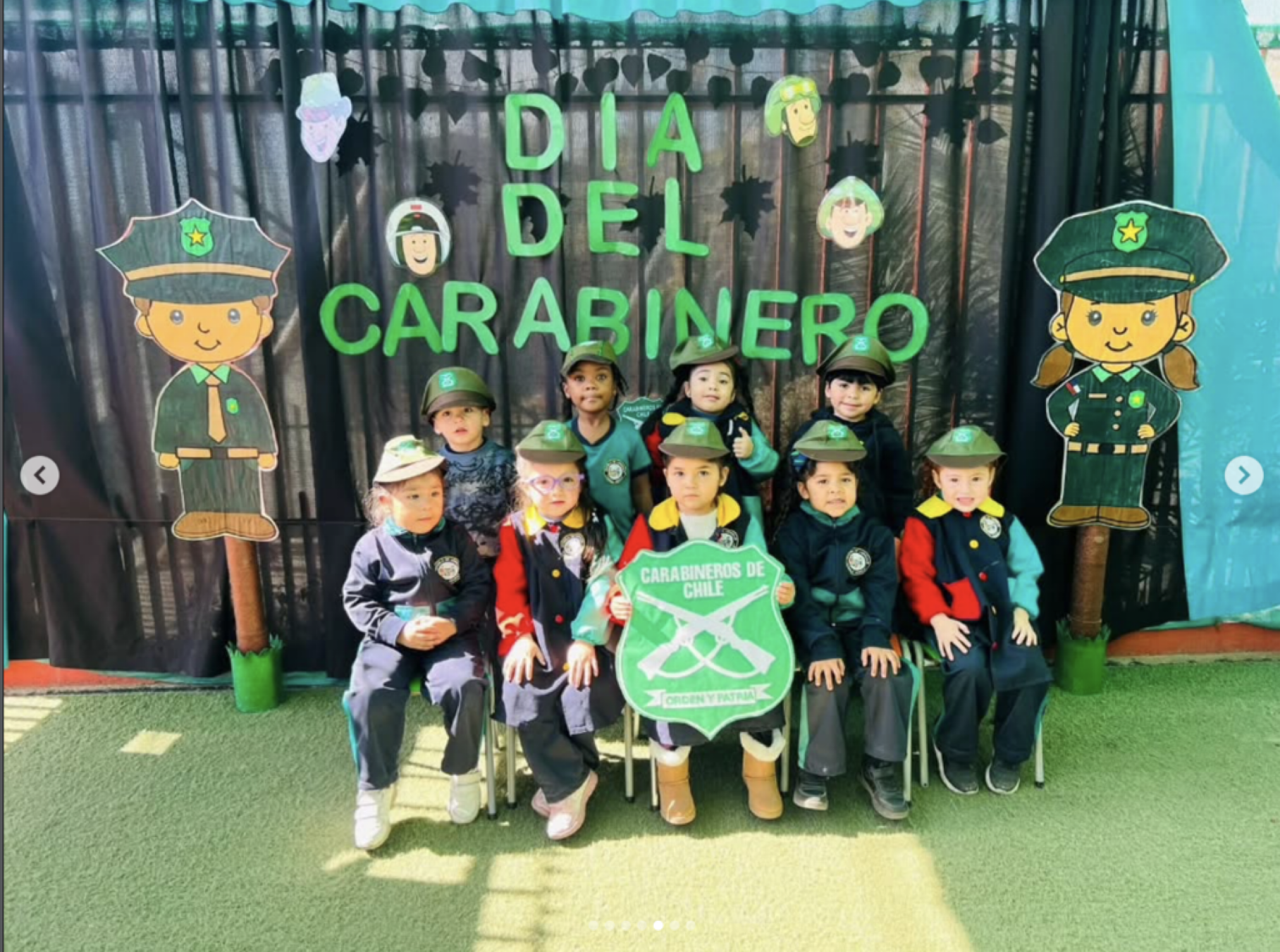 Grupo de niños vestidos con disfraces de carabineros de Chile en una celebración del día del carabinero, con decoraciones alusivas y un cartel que dice 'Día del Carabinero'.