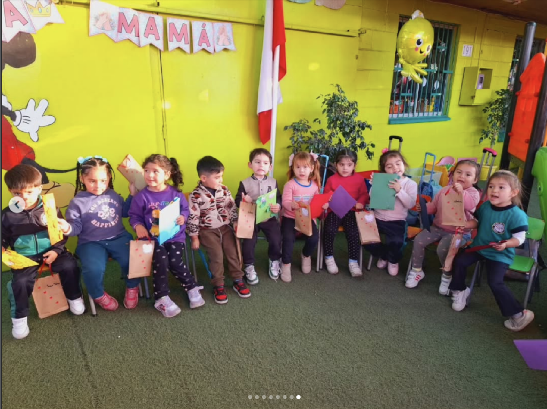 Niños y niñas sentados en una fila en una ceremonia o evento en un lugar decorado con globos y dibujos, con un fondo amarillo y una pared decorada con letras que dicen 'A MAMÁ'