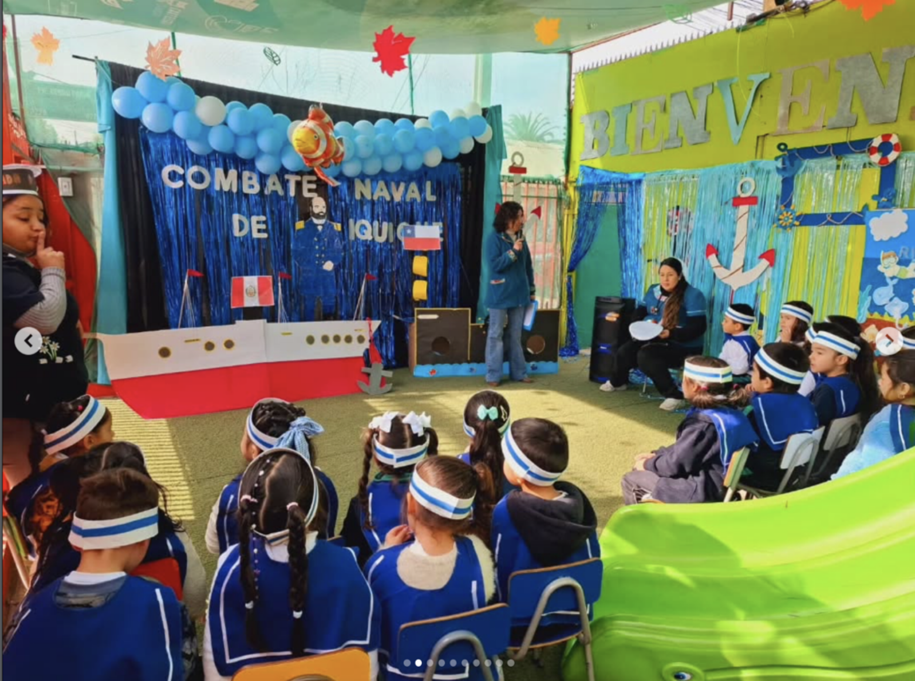 Niños sentados en una sala decorada con temática naval, escuchando a una mujer que habla, en un evento escolar con tema de combate naval y bienvenida.