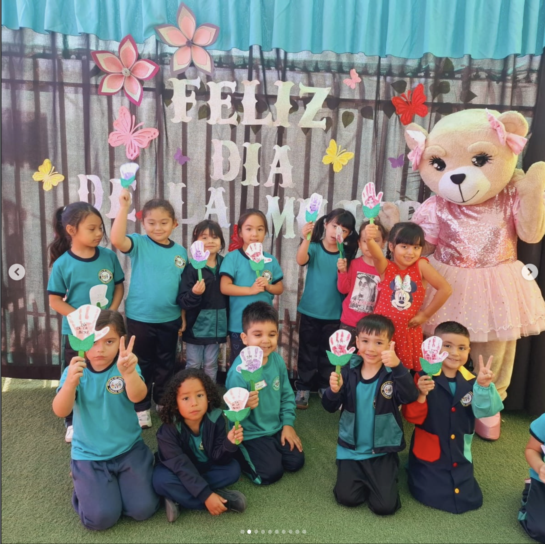 Niños en celebración del Día de la Madre en un evento escolar, sosteniendo manualidades de flores de papel, acompañados por una mascota gigante de oso con lazos rosados y una decoración con letras que dice 'FELIZ DÍA'.