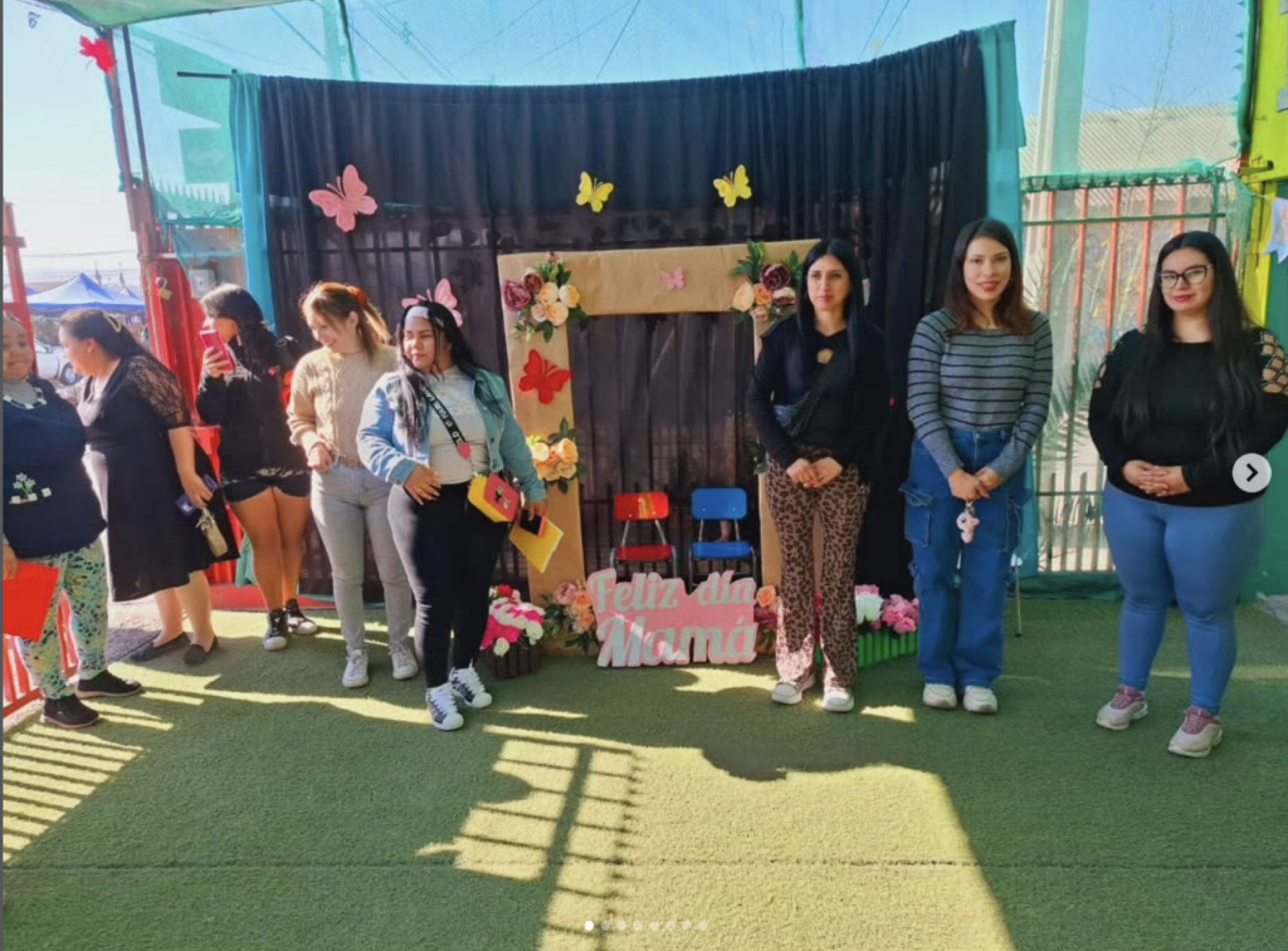 Grupo de mujeres y niñas celebrando en un evento con decoración floral y un cartel que dice "Feliz día Mamá". Muestran diferentes expresiones y están de pie frente a una estructura decorativa en un espacio abierto.