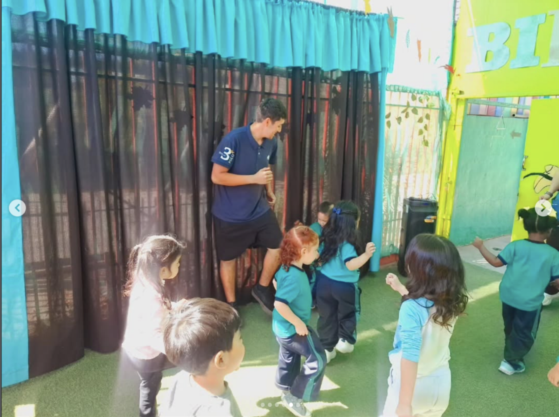 Niños de escuela en una actividad al aire libre, algunos están de pie y otros sentados, junto a un adulto que parece ser un instructor, en un espacio decorado con cortinas negras y paredes verdes y amarillas.