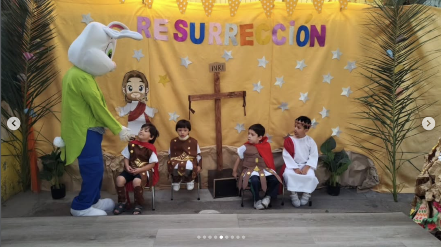 Niños representando la historia de la resurrección de Jesús en una obra escolar, con figuras infantiles y decoraciones temáticas, incluyendo una cruz, un texto que dice 'Resurrección' y varios personajes en vestuarios bíblicos.