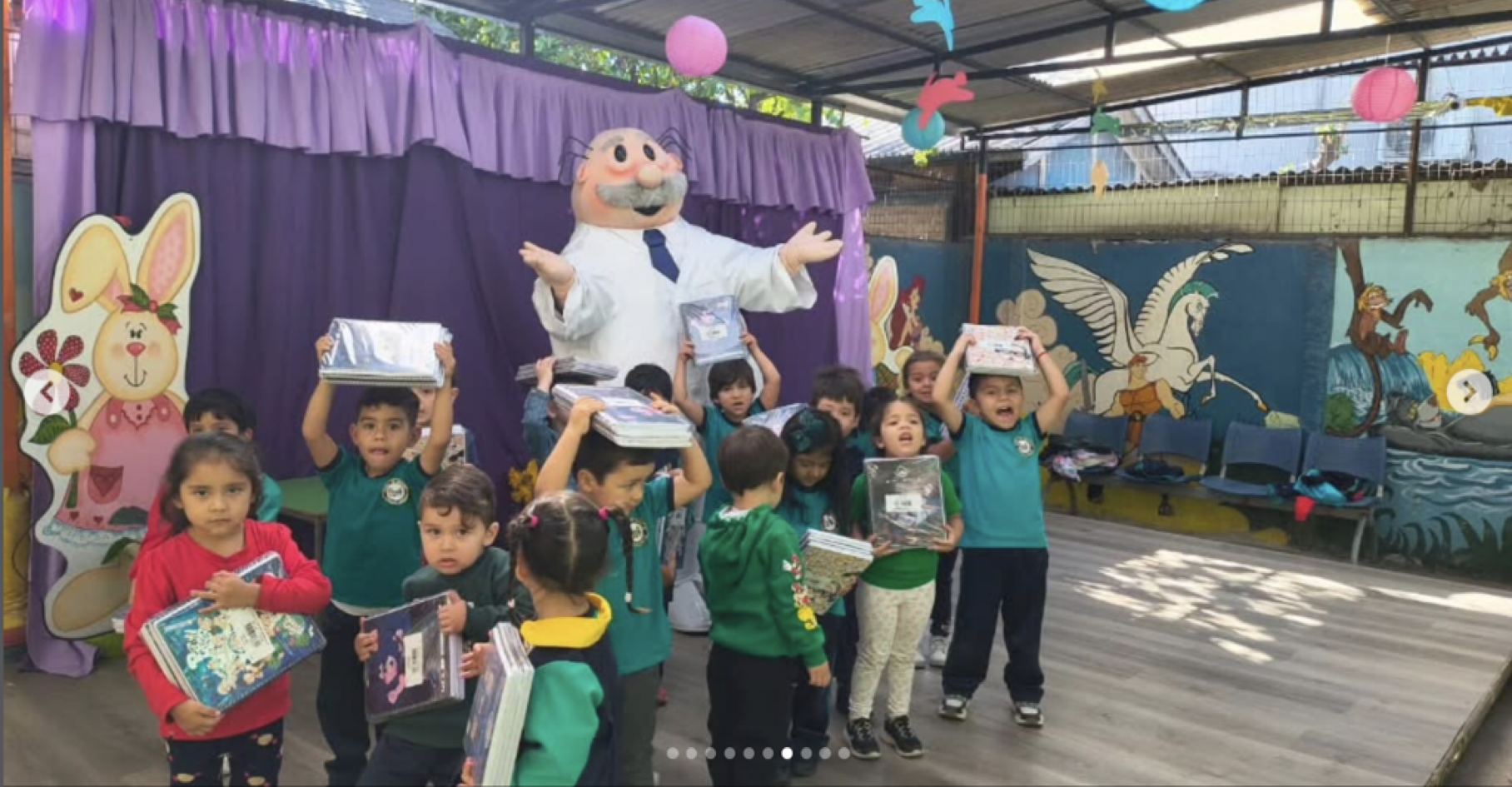 Grupo de niños en una celebración escolar con decoraciones coloridas, algunos sosteniendo libros y otros levantando pilas de libros, en un escenario decorado con un conejo gigante y personajes de dibujos animados.
