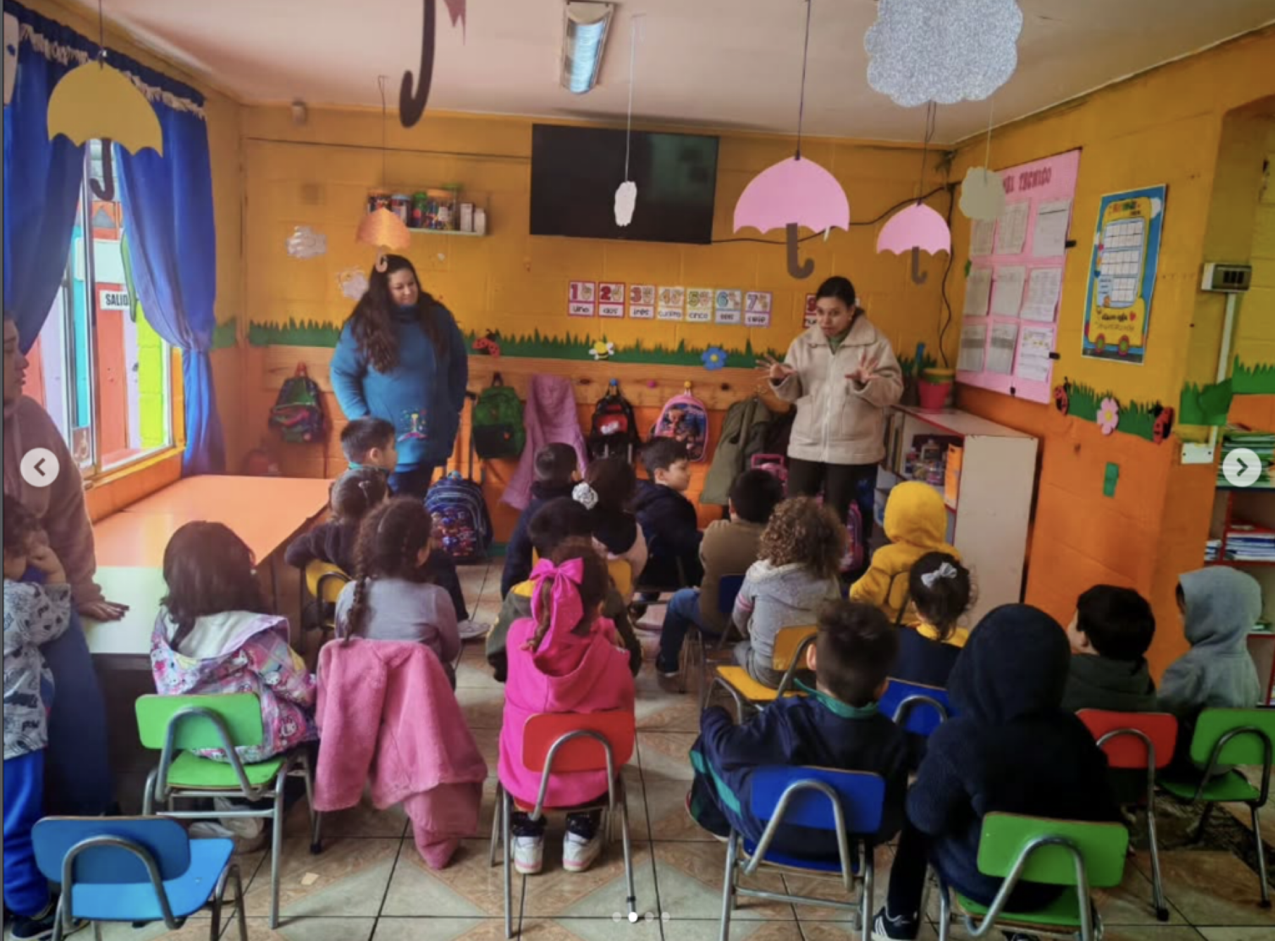 Niños en una aula escolar escuchando a una maestra en la frente de la clase. La habitación está decorada con dibujos de paraguas y otros objetos coloridos en las paredes.