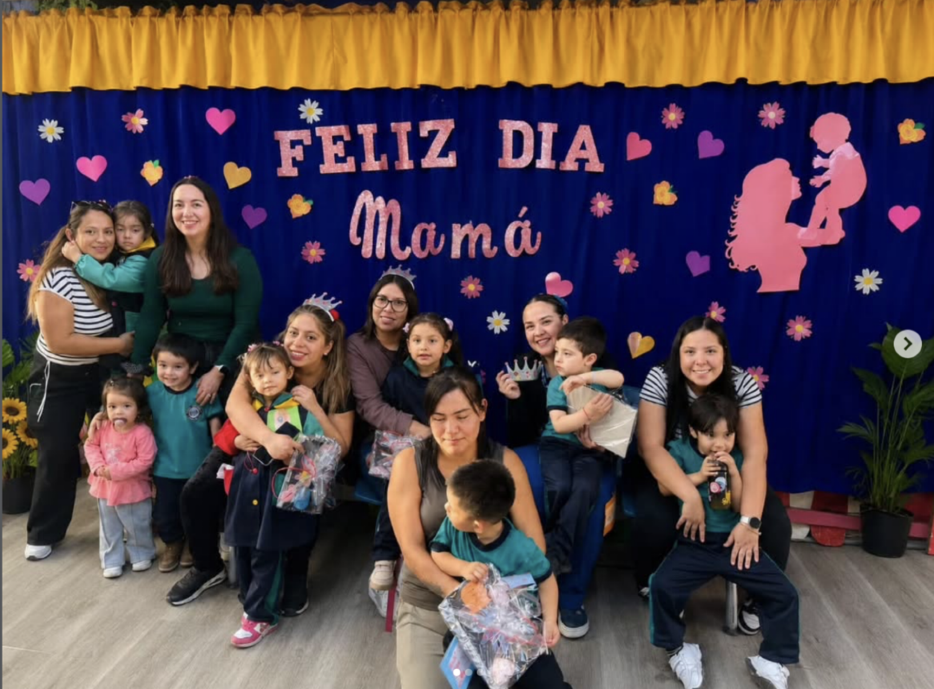 Grupo de personas en una celebración del Día de la Madre, con un cartel que dice 'Feliz Día Mamá', decoraciones con corazones, flores y una silueta de madre con un niño en brazos en el fondo.