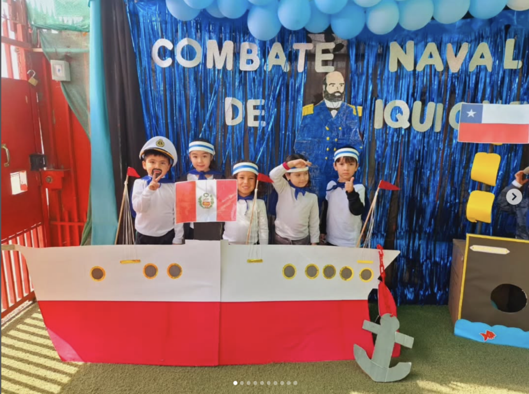 Niños disfrazados de marineros en un escenario decorado con un tema naval, con un cartel que dice "Combate Naval de Aquí" y una bandera peruana, simulando un barco y el mar.