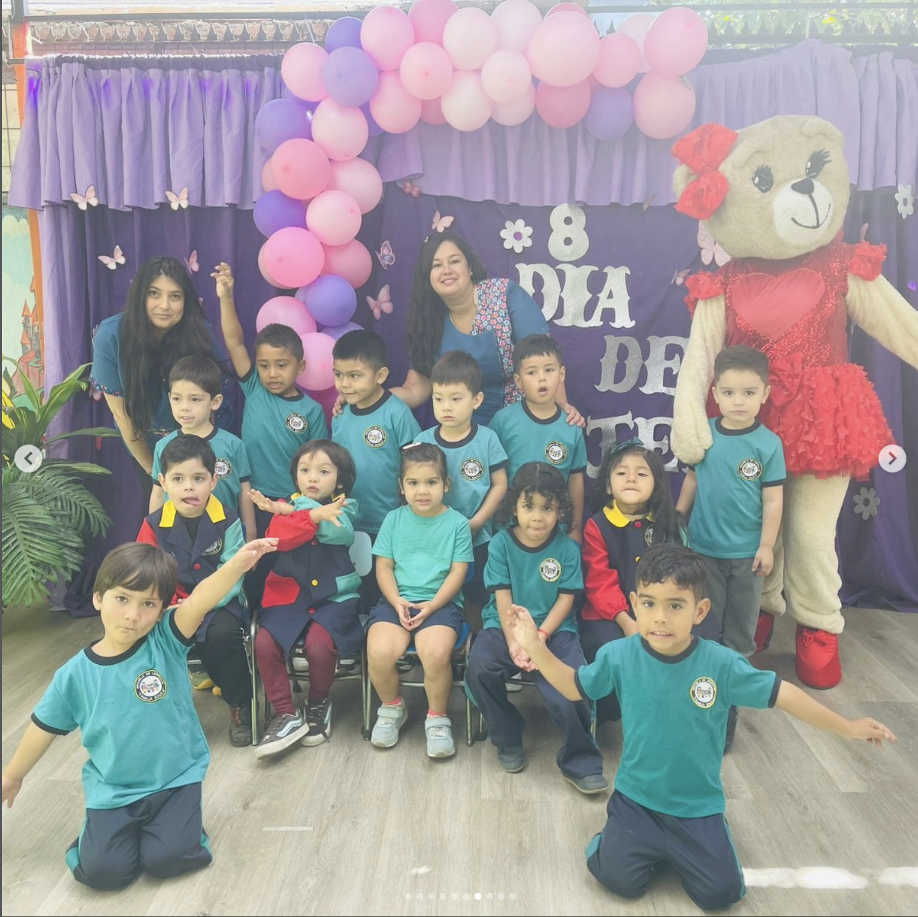 Niños y niñas con camisetas azules posando con maestras y un peluche gigante en una decoración de fiesta con globos color rosa y lila, y fondo de cortinas moradas con mariposas y números de cumpleaños.