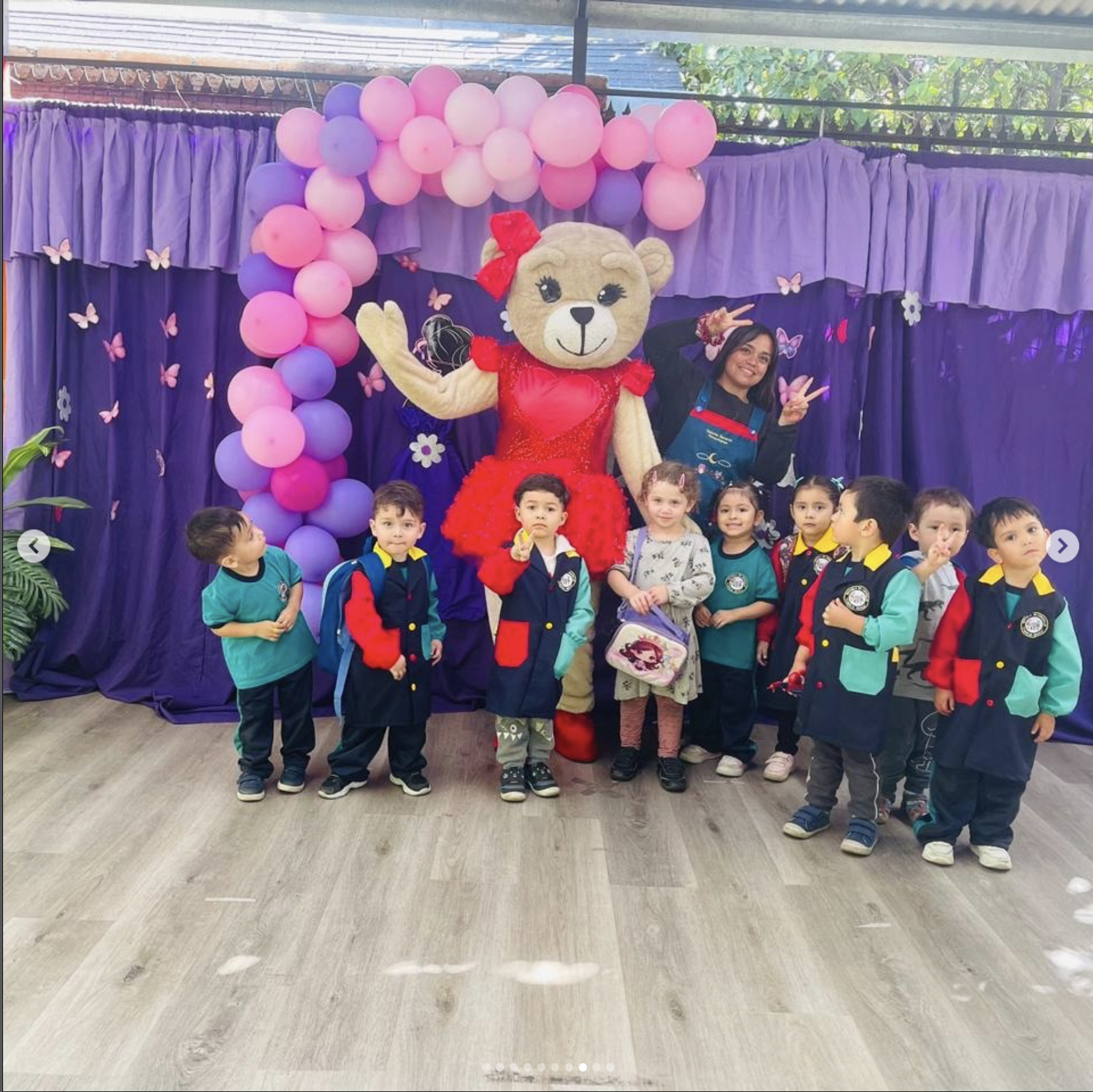 Niños en una celebración escolar con una mascota de león vestida de vestido rojo y muchas globos de colores en el fondo.